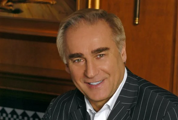 A smiling middle-aged man in a pinstripe suit with a white shirt sitting indoors with wood paneling behind him.