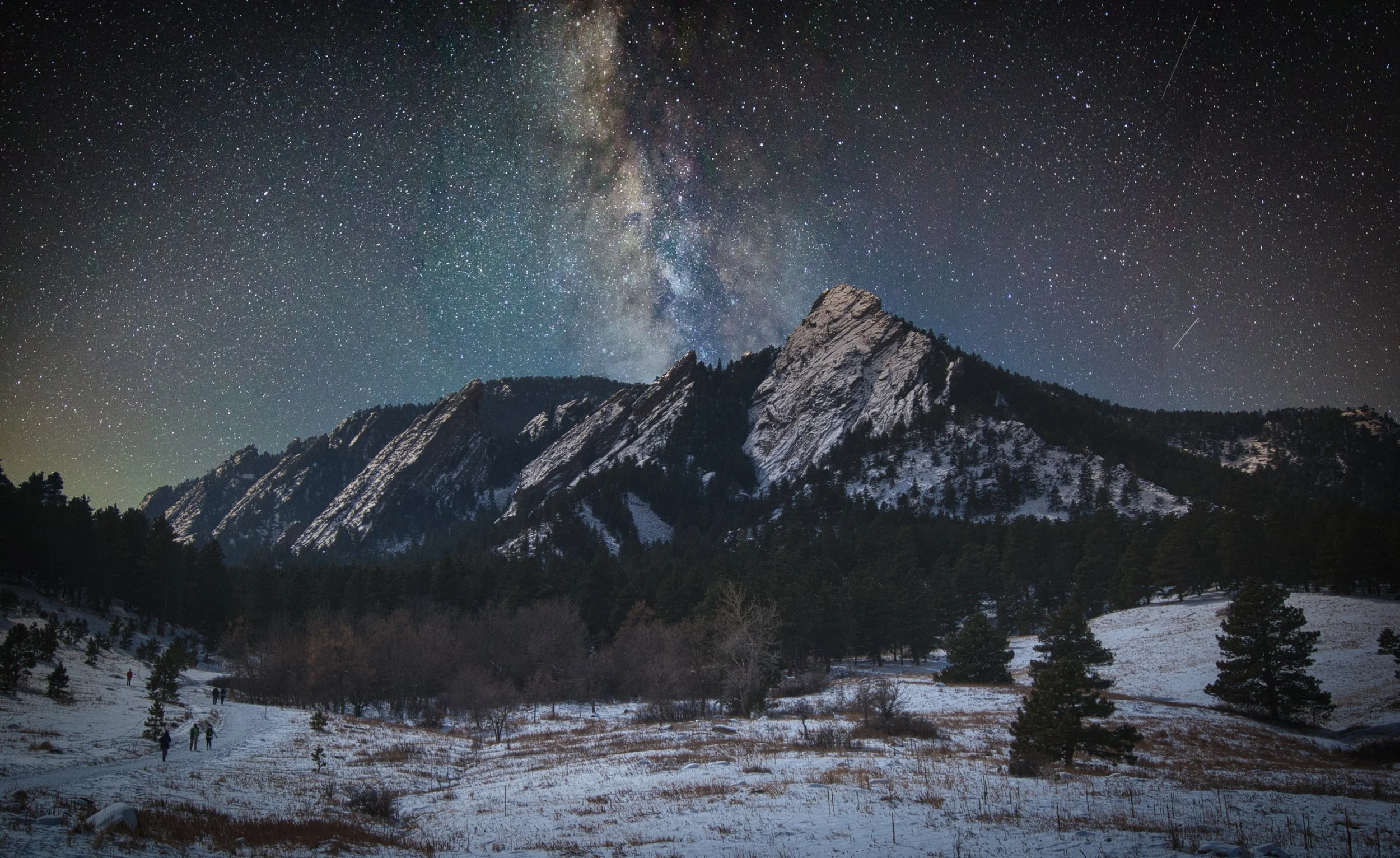 Nighttime landscape of snow-covered mountains with a star-filled sky and the Milky Way galaxy visible overhead.