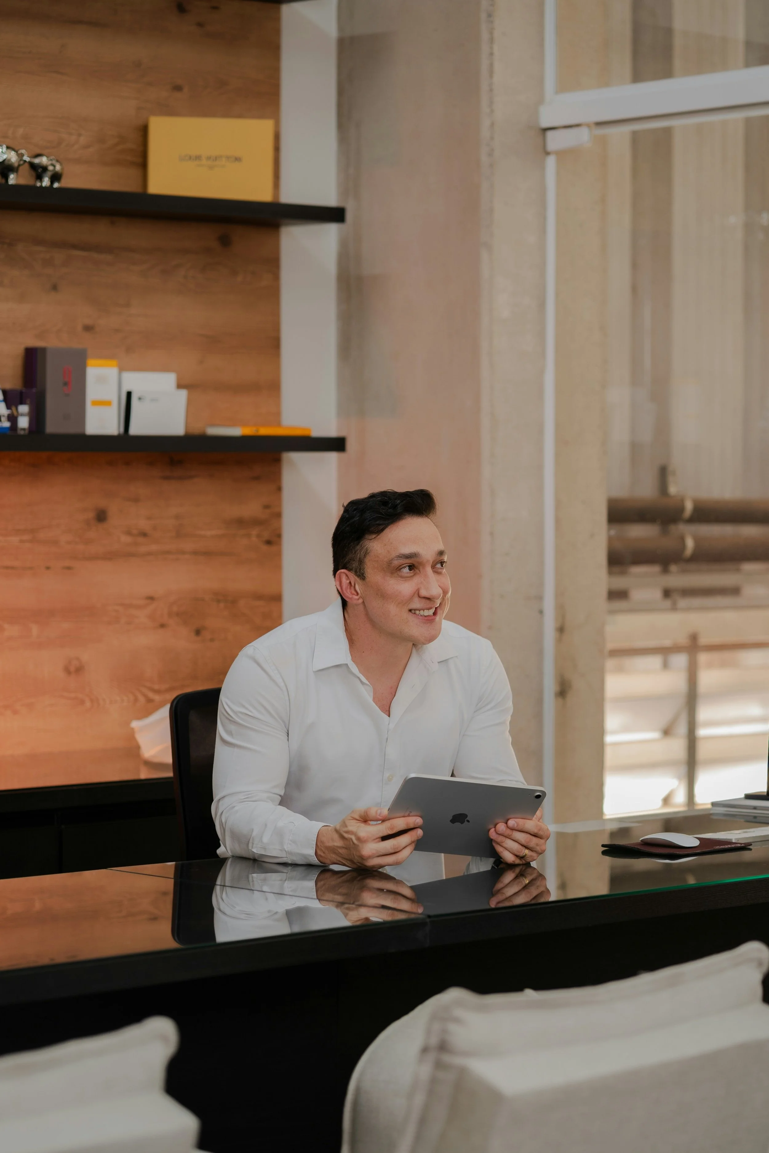 A smiling man in a white blouse is holding an ipad and sitting on a desk in a nice office