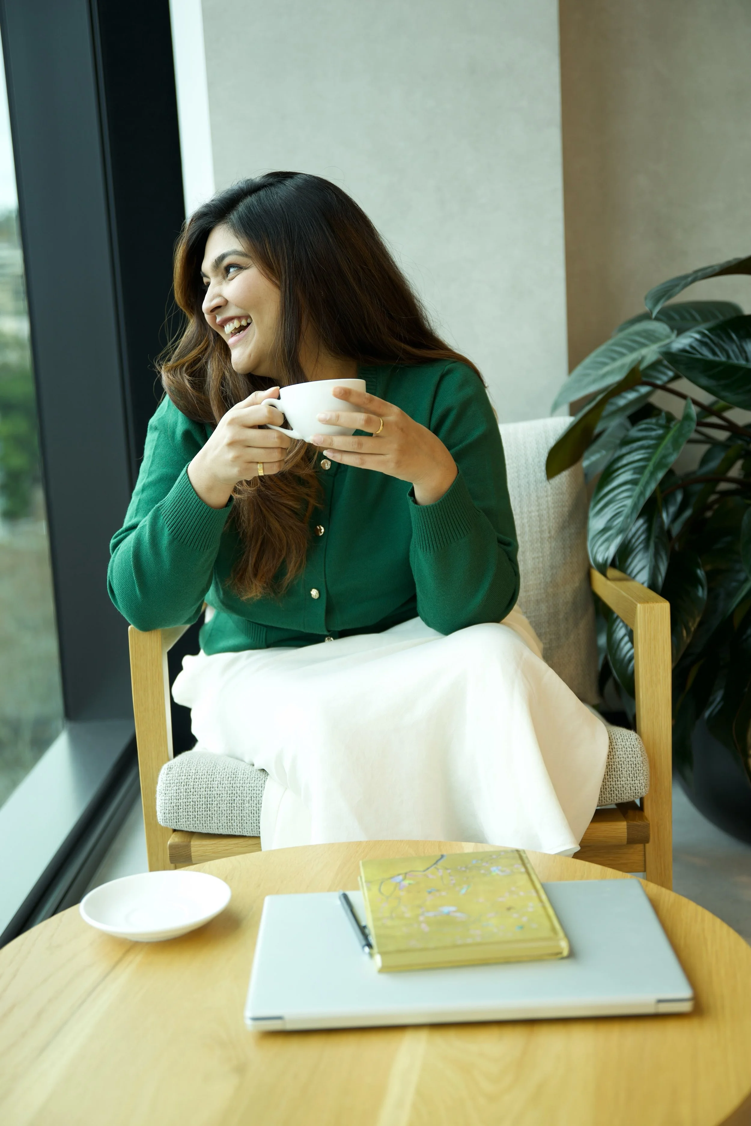 A woman sitting on a chair near a window, smiling and holding a white mug, with a table in front of her that has a closed yellow and decorated books, a closed white laptop, and a white plate.