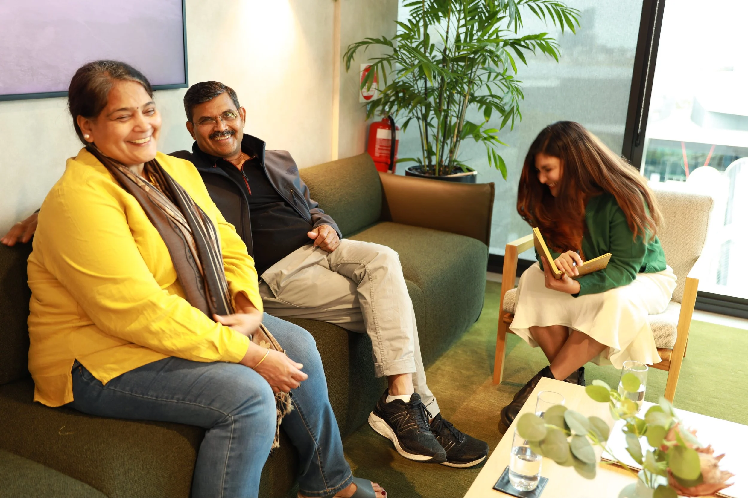 Four people sitting on a green couch in a modern office lounge, smiling and enjoying a relaxed moment. A woman wearing a yellow jacket and scarf is on the left, next to a man with glasses and a dark jacket. A young woman with long hair and a green sweater is sitting on a chair, holding a tablet and laughing. There are drinks on a table in front of them and a large plant in the background near a window.