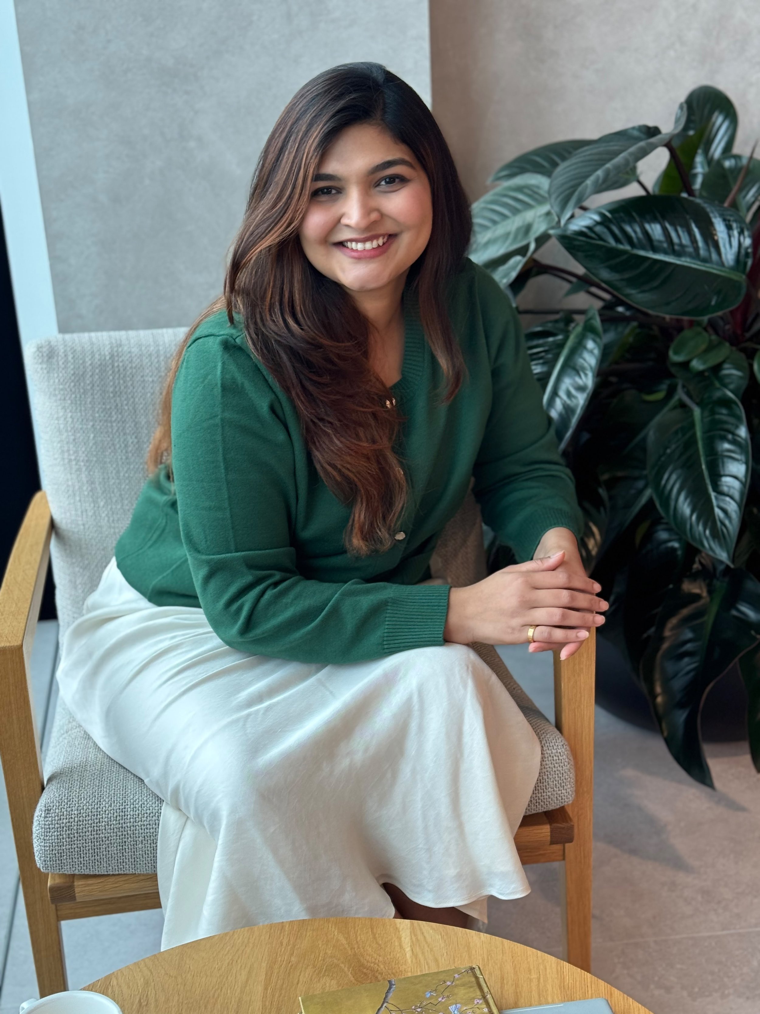 A smiling woman with long brown hair sitting on a wooden chair with a cushioned seat, wearing a dark green sweater and a light-colored skirt, with greenery in the background.