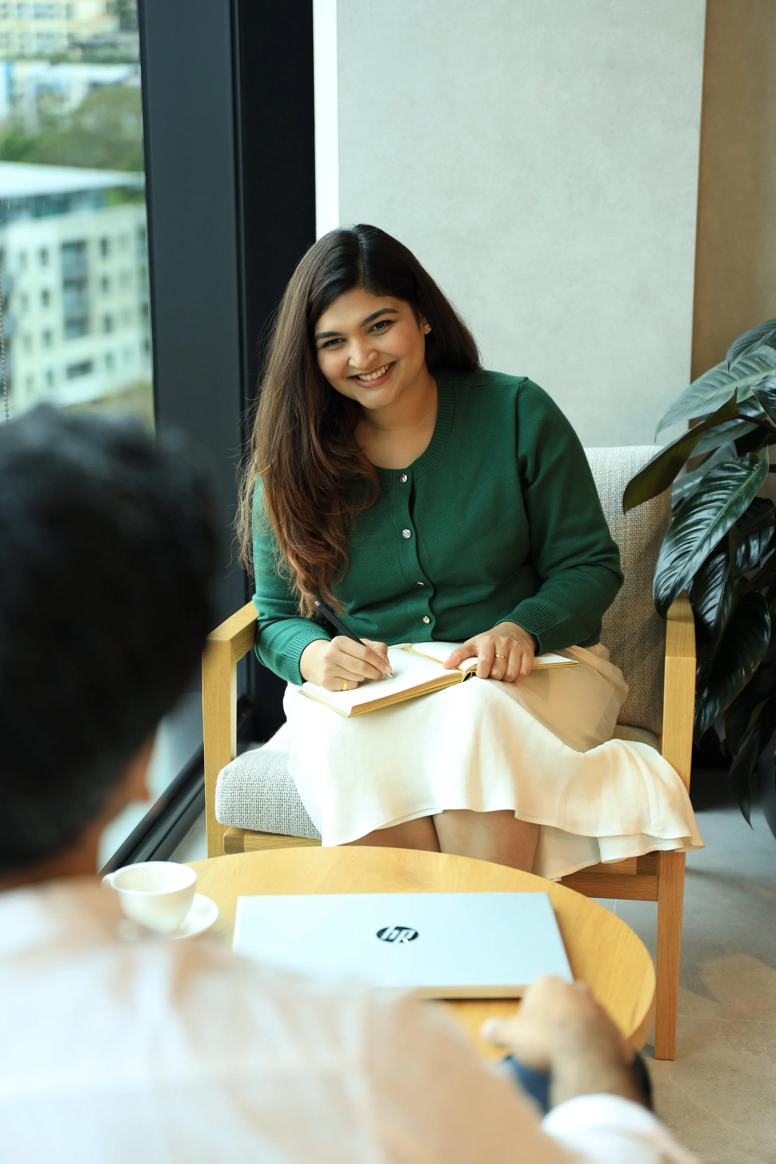 A woman with long dark hair smiling while sitting in a chair during a meeting, writing in a notebook. She is wearing a green cardigan and a beige skirt, and there is a large window behind her with city buildings visible outside. There is a plant to her right and a laptop and a cup on the table in front of her.