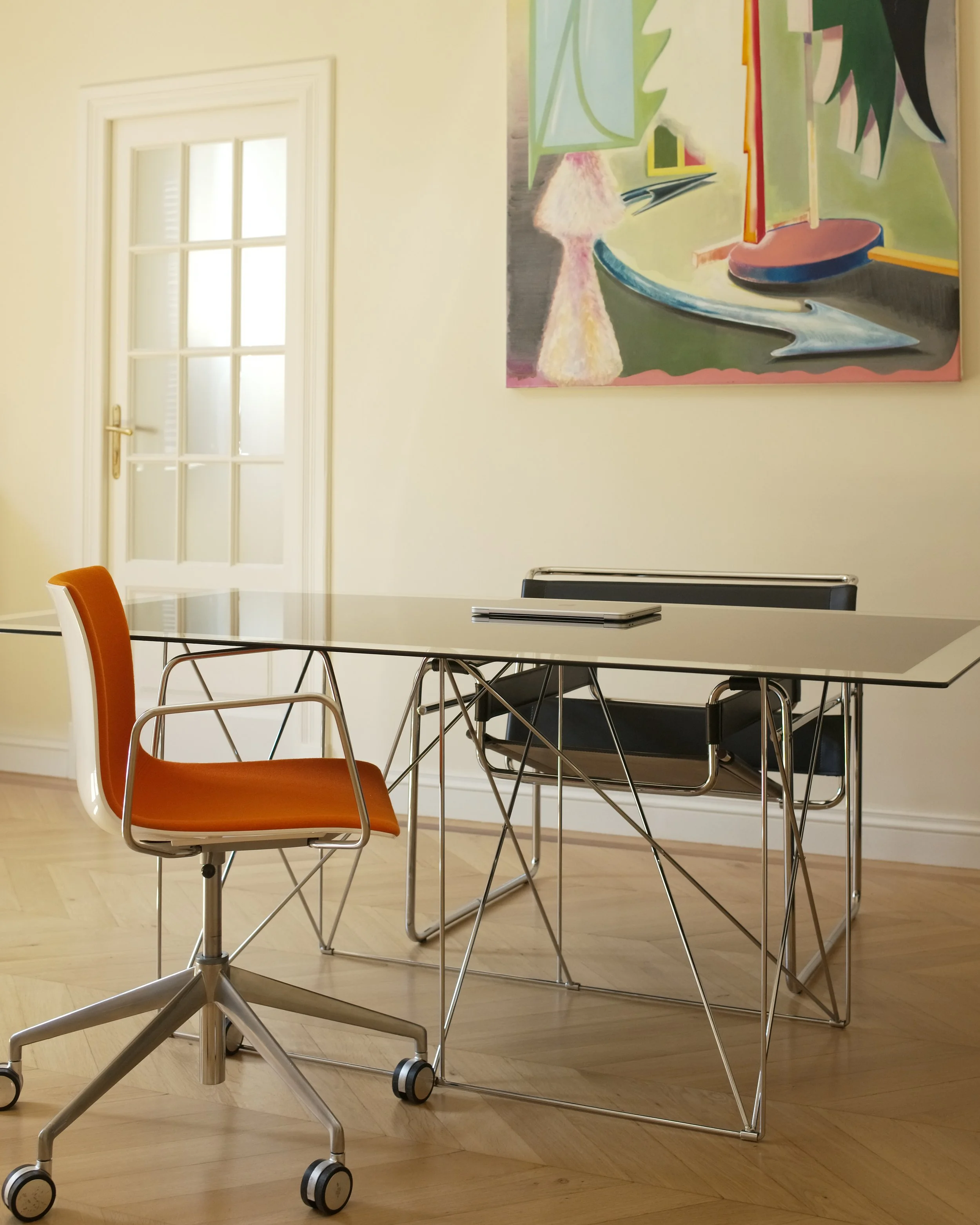 An interior room with a white door, colorful abstract painting, table with a laptop, orange chair, and a hospital wheelchair in the foreground.