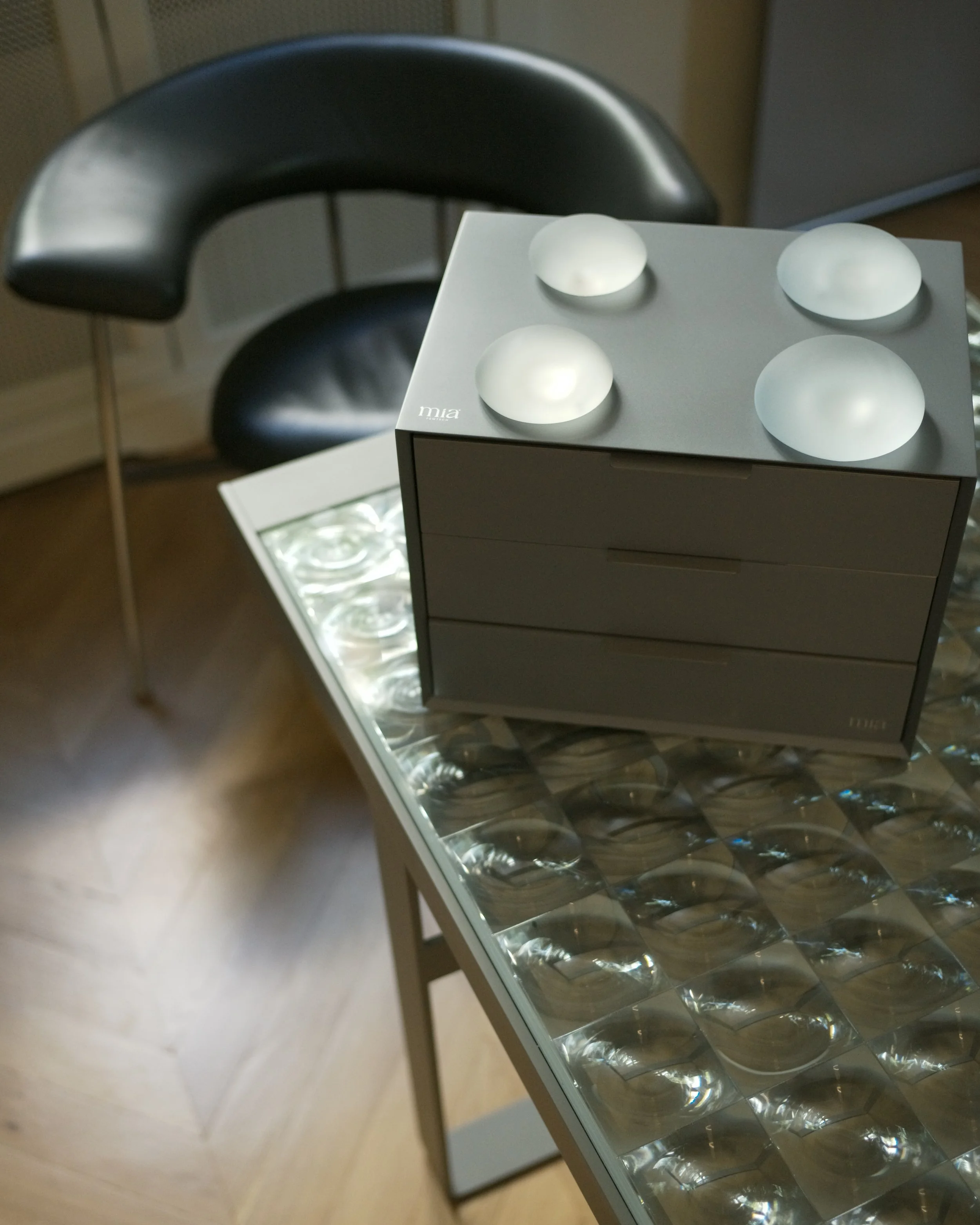 A black wheelchair with a curved armrest visible behind a small table with a silver box on top. The box contains four white, dome-shaped objects. There is a reflective surface underneath the table.