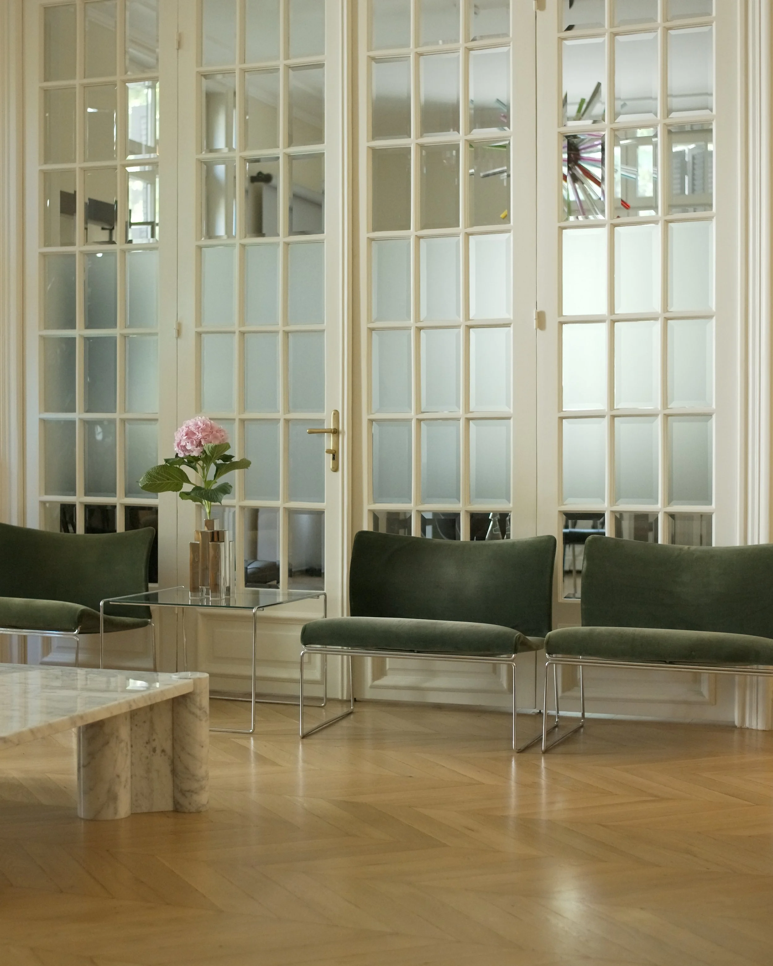 Interior of a room with large glass-paned doors, a marble-topped table, a pink hydrangea in a vase, and green chairs.