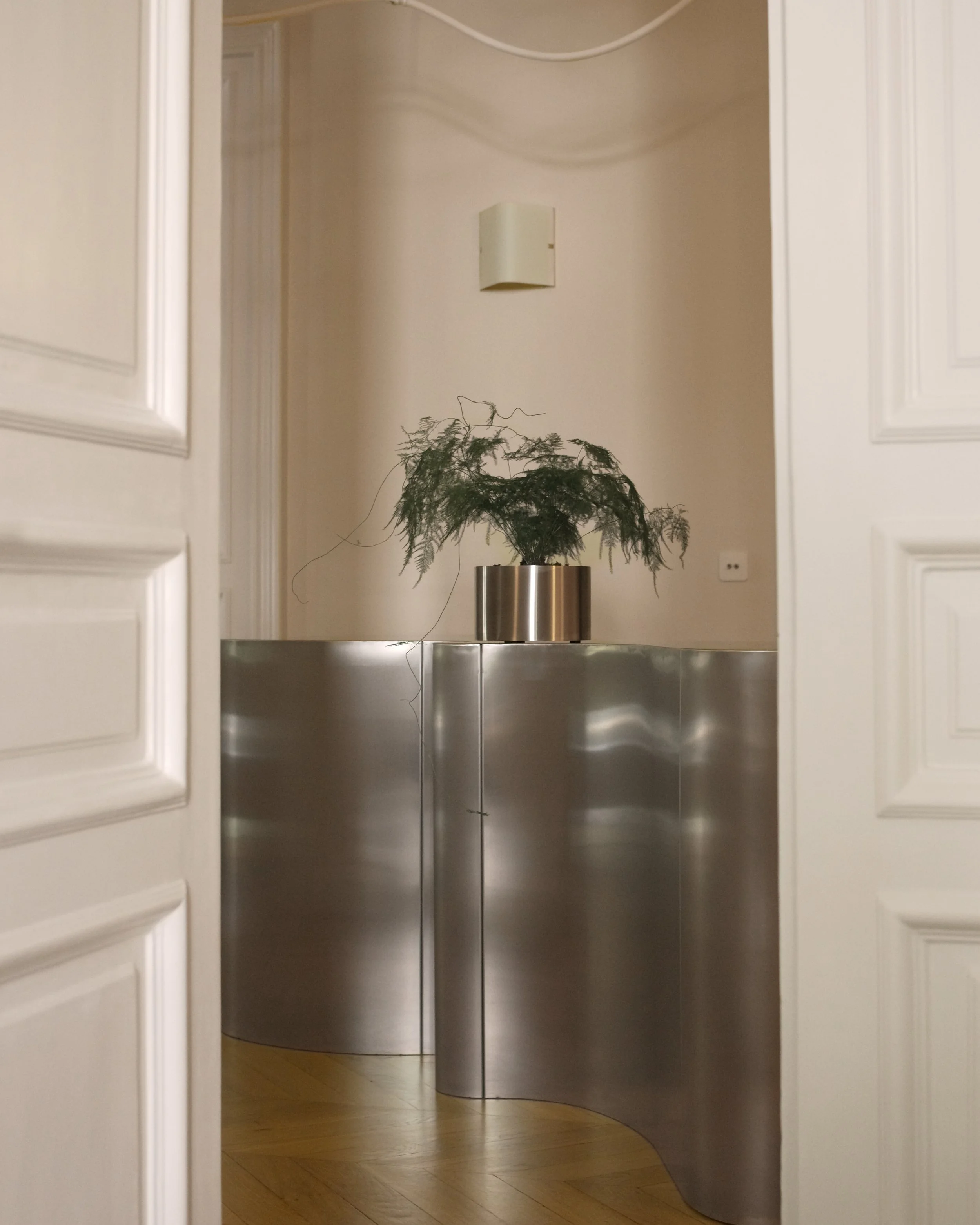 A modern interior space with a curved stainless steel wall and wooden flooring, viewed through a doorway with white trim.