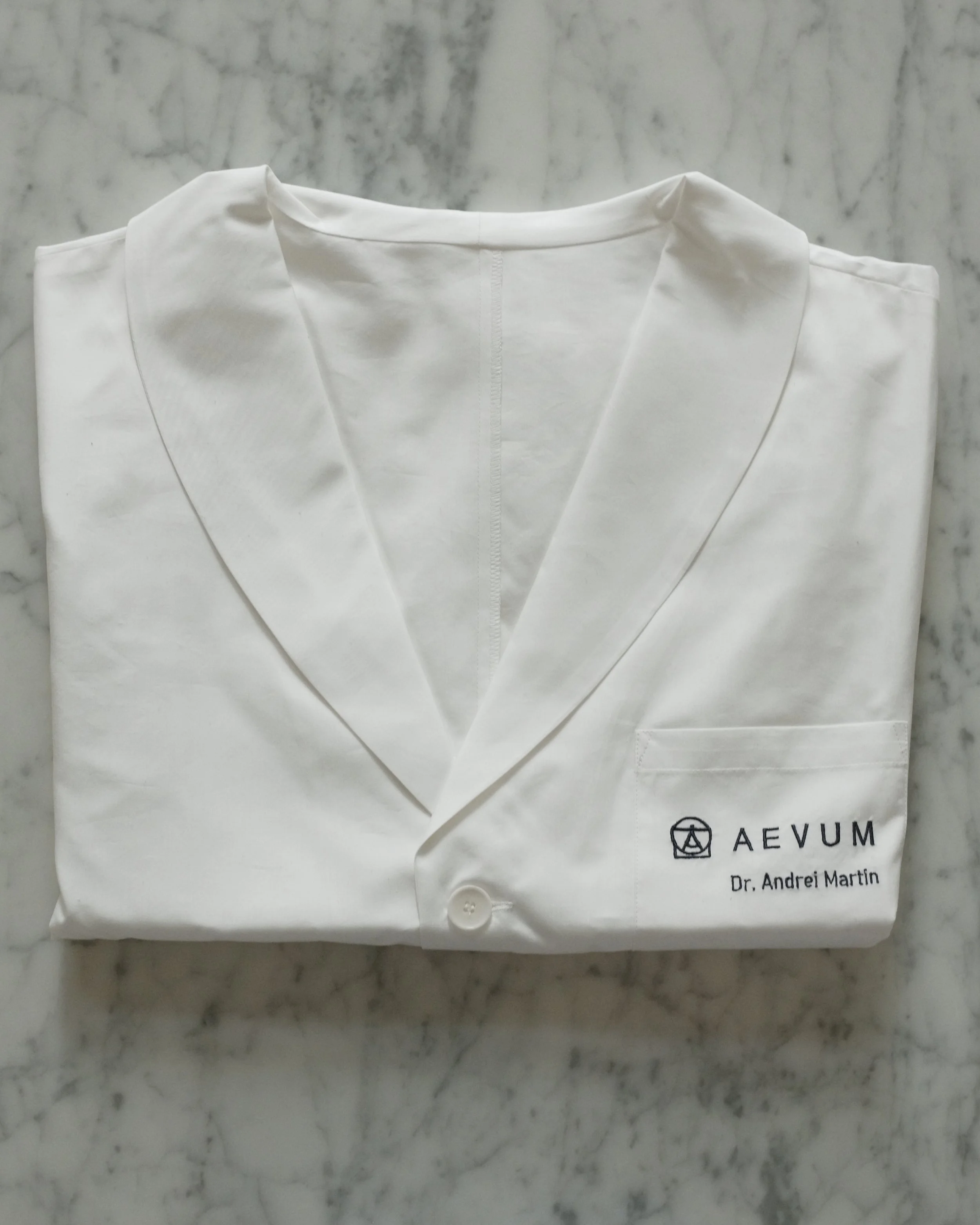 White folded lab coat or medical coat with logo and text on the right bottom corner placed on a marble surface.
