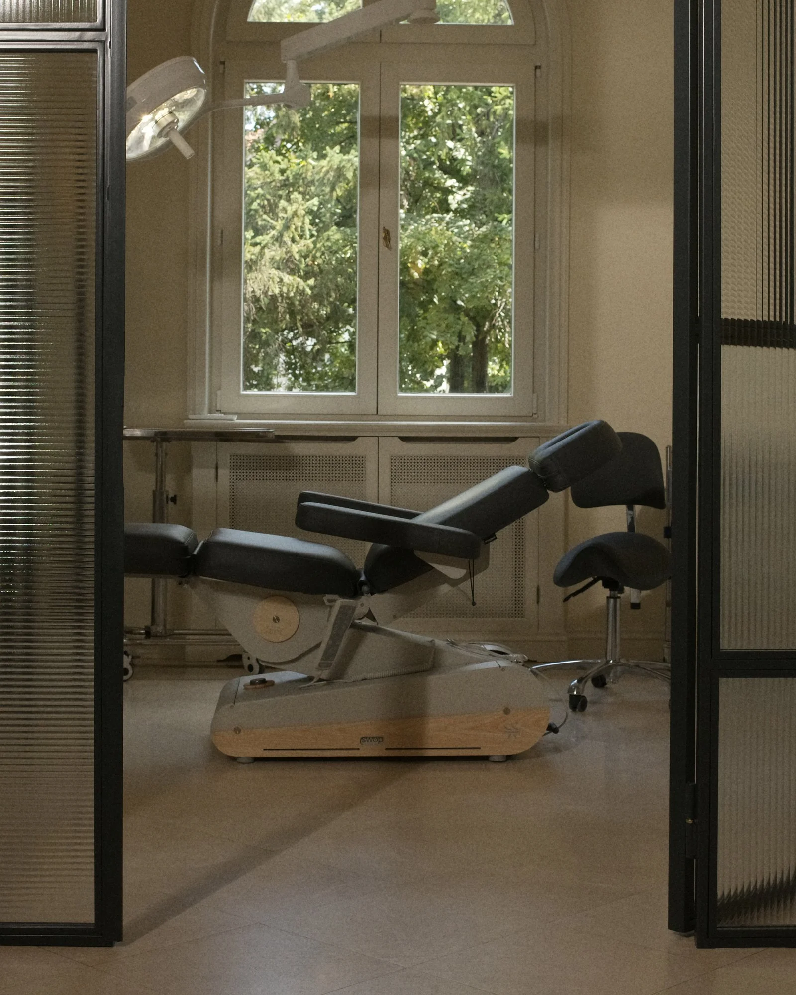 A hospital or medical examination room with a medical chair, a large window with green trees outside, an overhead examination light, and privacy screens on either side.