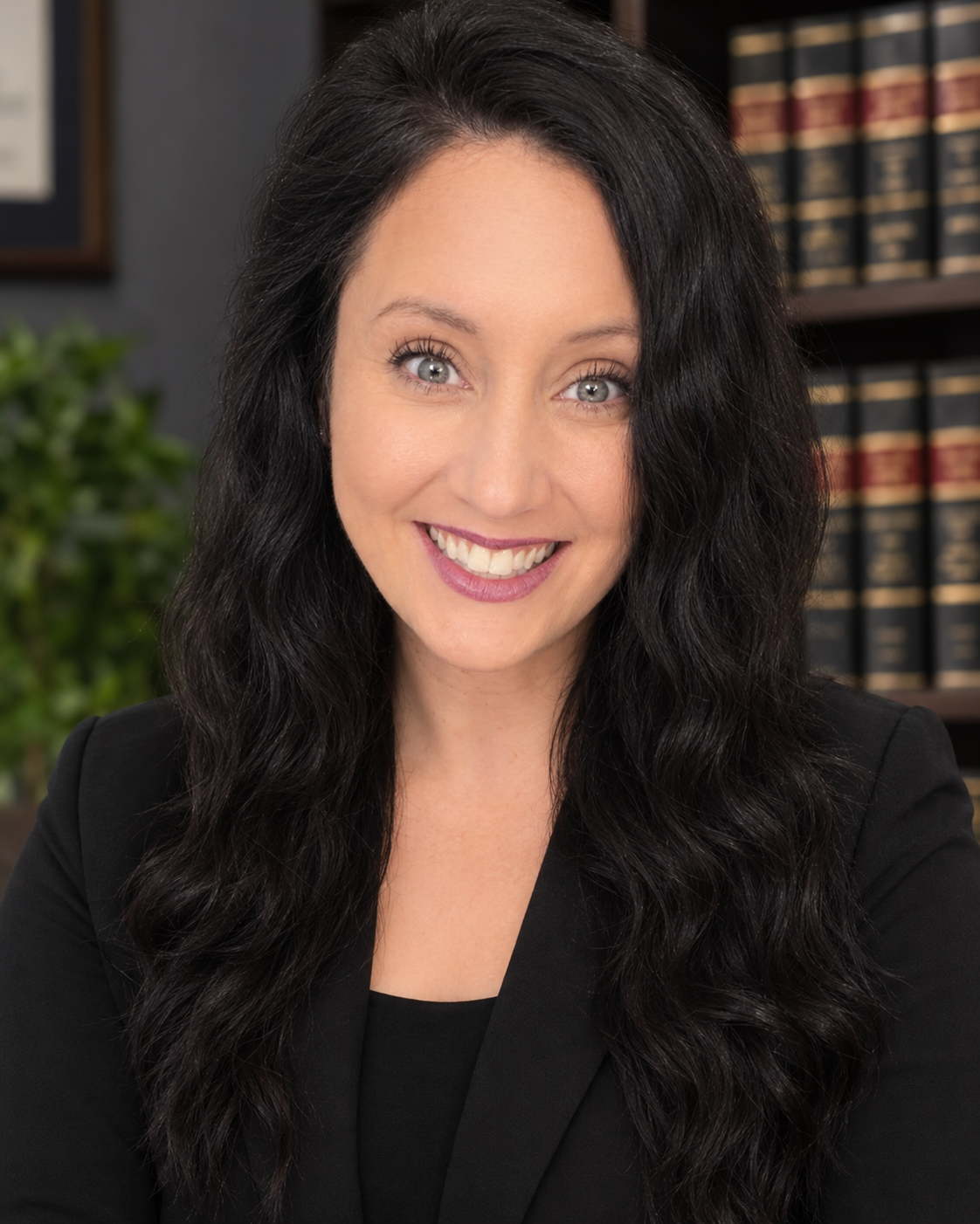 A woman with long dark hair and blue eyes smiling, dressed in a black blazer, in an office setting with a bookshelf and a potted plant in the background.