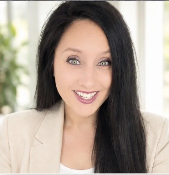 Portrait of a smiling woman with long black hair wearing a beige blazer in a bright indoor setting.