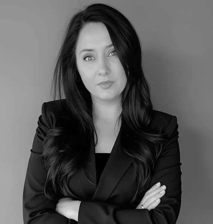 A woman with long dark hair and light eyes, wearing a black blazer, standing with her arms crossed against a plain background.