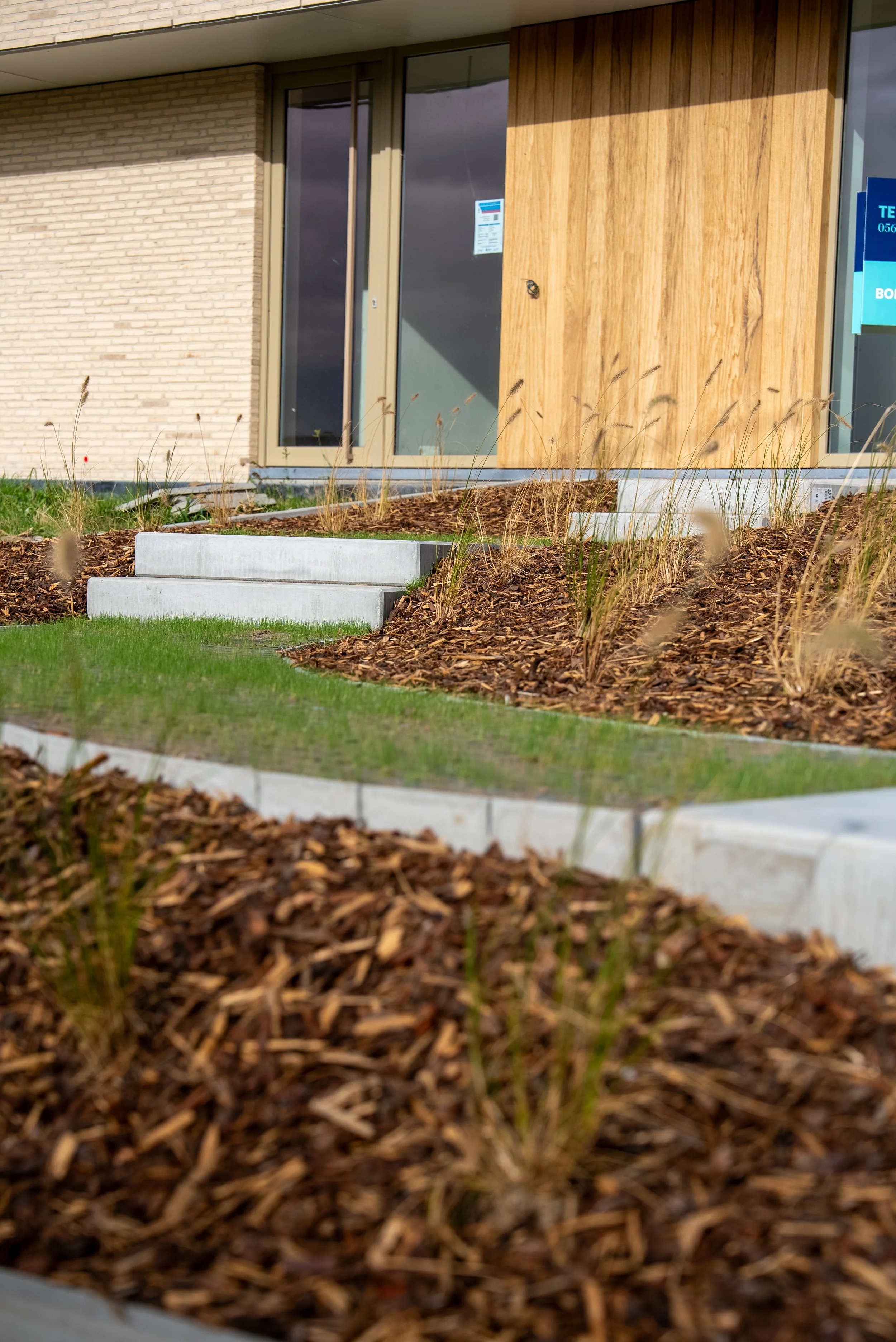 De afbeelding toont de gevel van een modern huis met grote ramen, houten en stenen gevels, een trap, en een tuin met droog gras en planten.