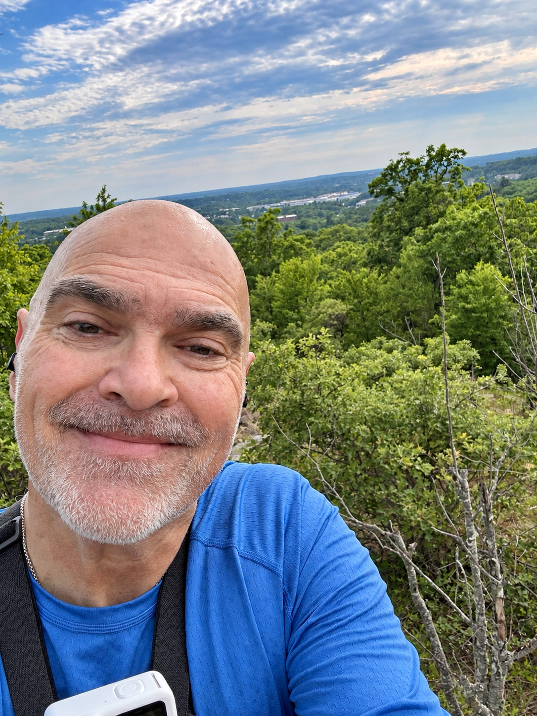 Bob Catinazzo taking a selfie at a scenic overlook with trees and a distant skyline behind him.