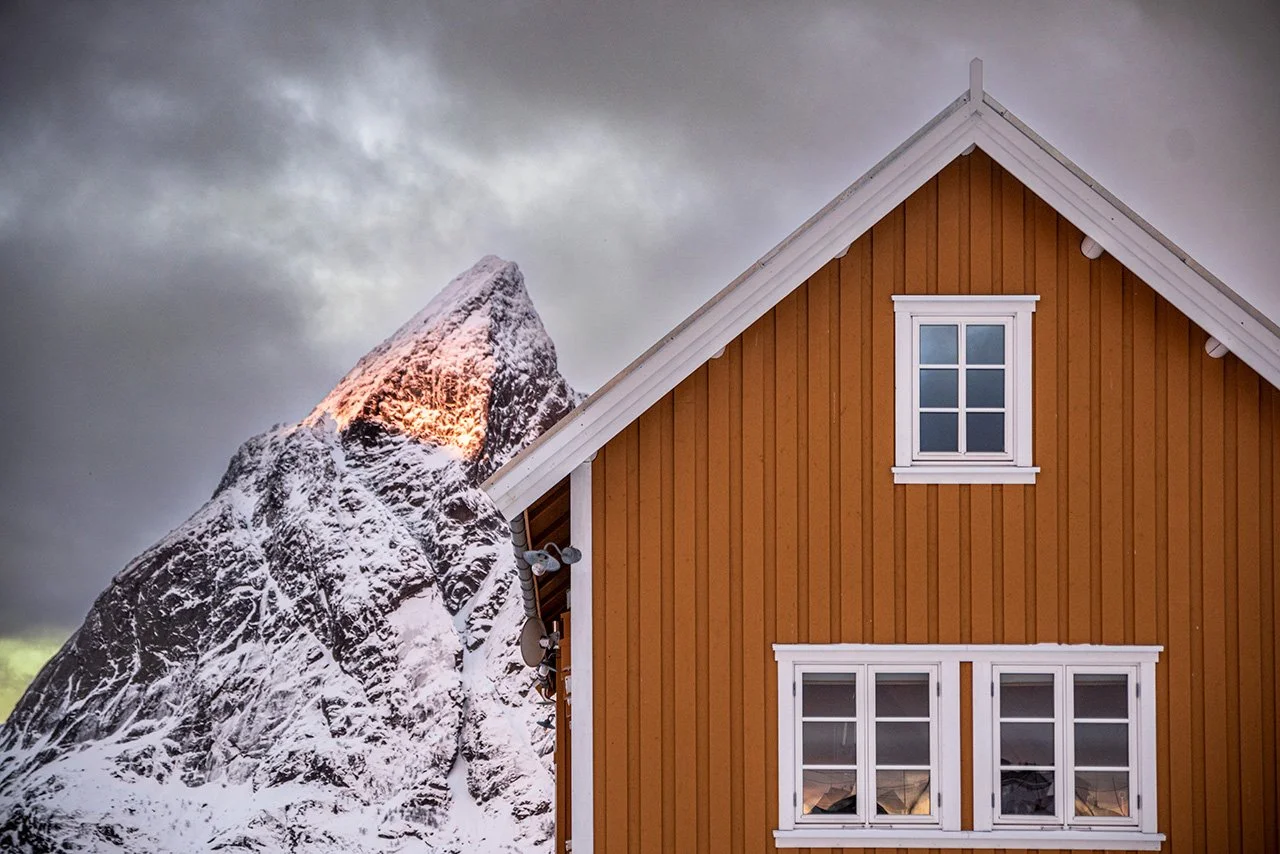 A house with orange wooden siding and white-framed windows next to a snow-covered mountain peak under cloudy skies.