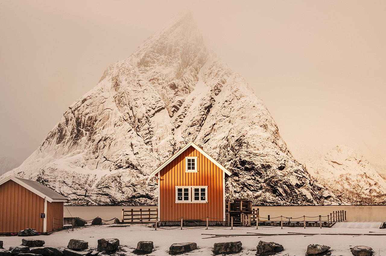 A small orange wooden house with white windows near a large snow-covered mountain, with snow on the ground and rocks in the foreground.