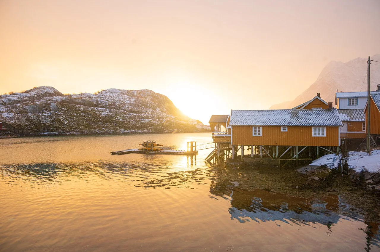 Sunset over a fjord with mountains and orange wooden houses on stilts by the water.