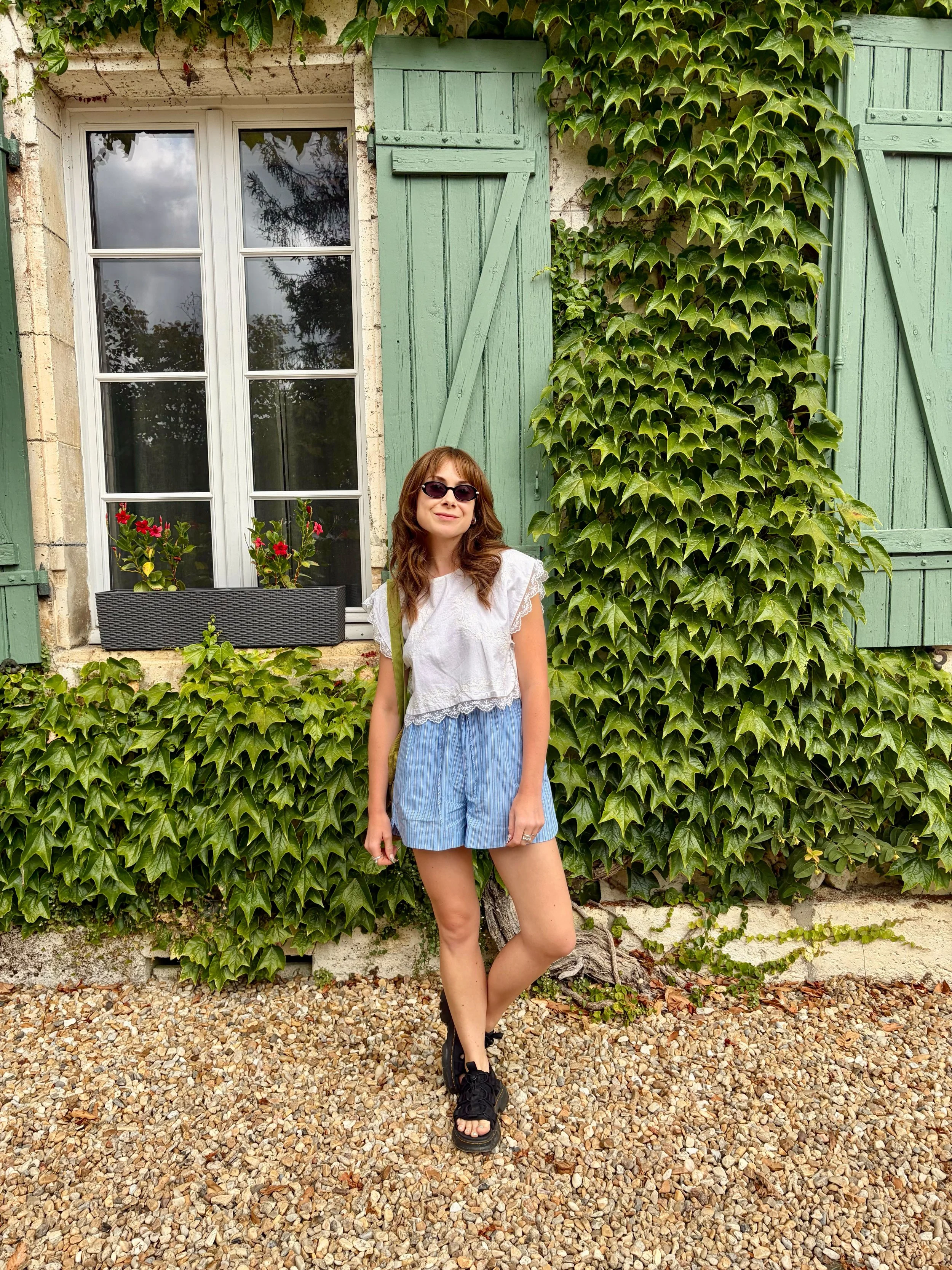 A woman with brown hair wearing sunglasses, a white top, and blue striped shorts standing in front of a house with green window shutters and ivy covering part of the wall. She is standing on a gravel ground.