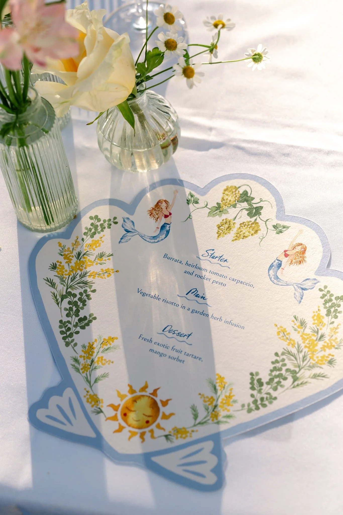 A table with glass vases containing flowers and a colorful menu with illustrations of a mermaid, sun, and floral designs.