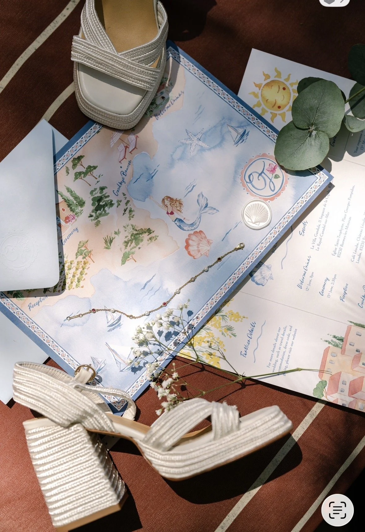 South of France wedding invite with a watercolor map background, white high-heeled shoes, a delicate bracelet, flowers, and printed event details on paper. The setup includes greenery and a warm-toned table surface.