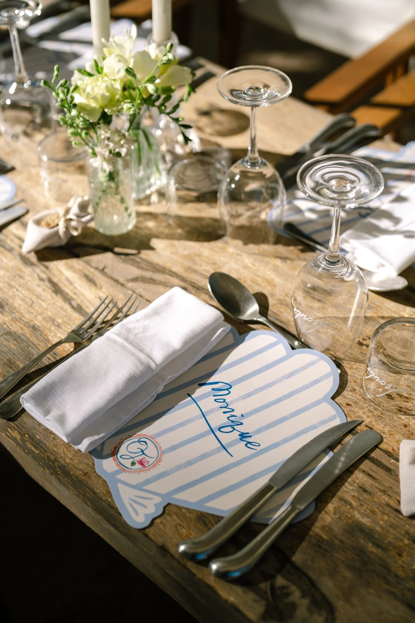 A rustic wooden table set for a meal with a white napkin, silverware, wine glasses, and a bouquet of white flowers in a glass jar. Featuring personalised shell menus.