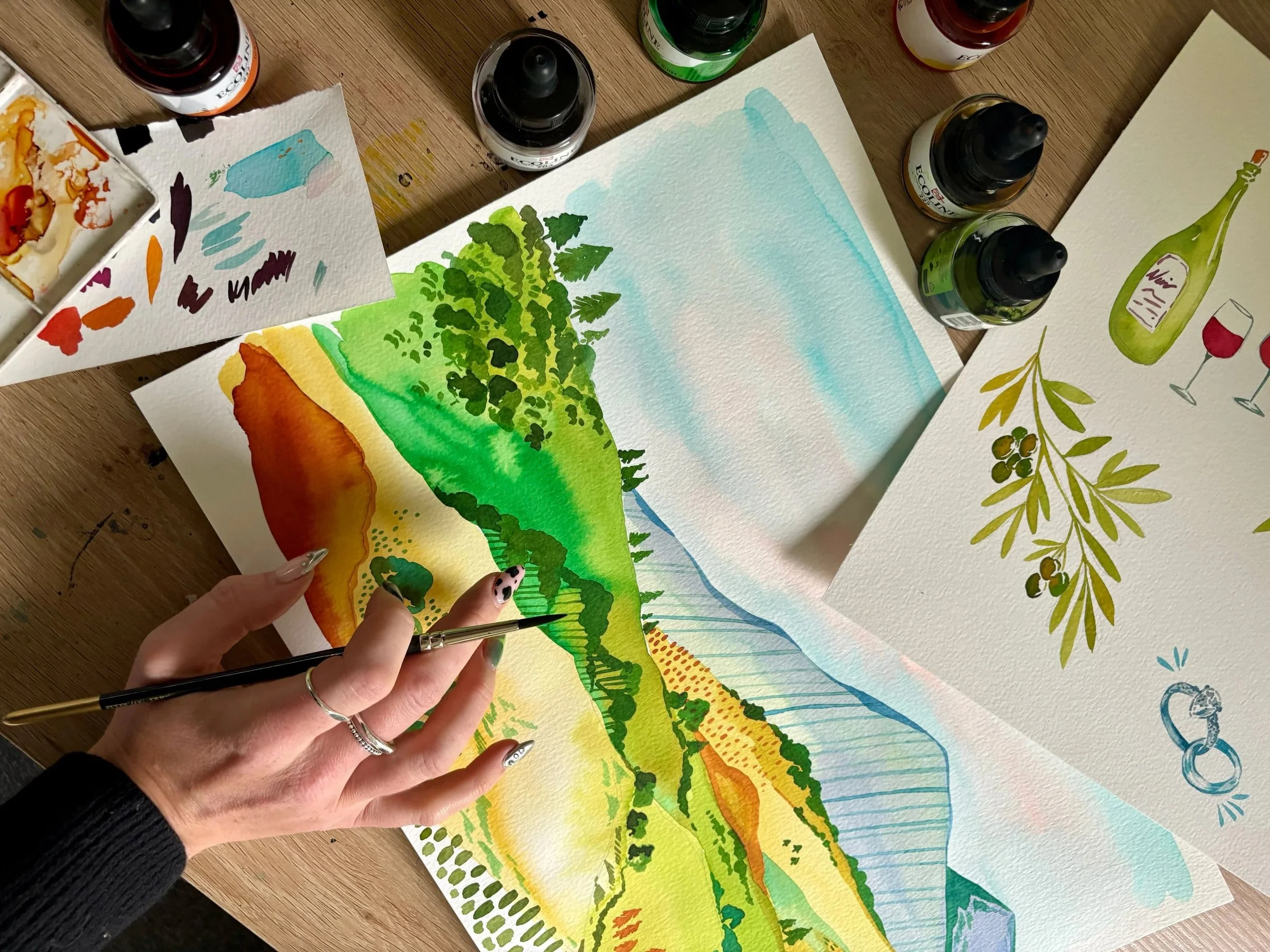 Hand painted illustration of hills in Tuscany. Various watercolor paintings and bottles of paint are spread on a wooden table.