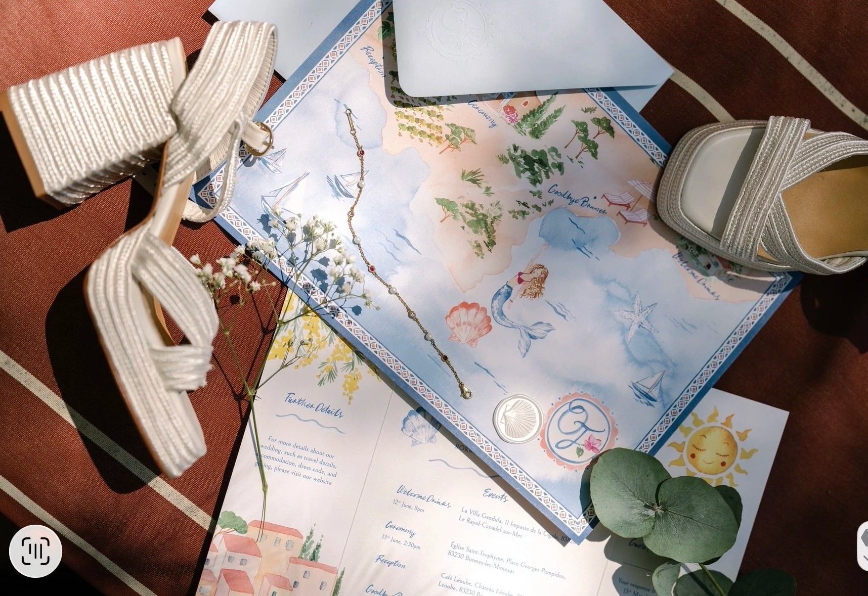 South of France wedding invite with a watercolor map background, white high-heeled shoes, a delicate bracelet, flowers, and printed event details on paper. The setup includes greenery and a warm-toned table surface.