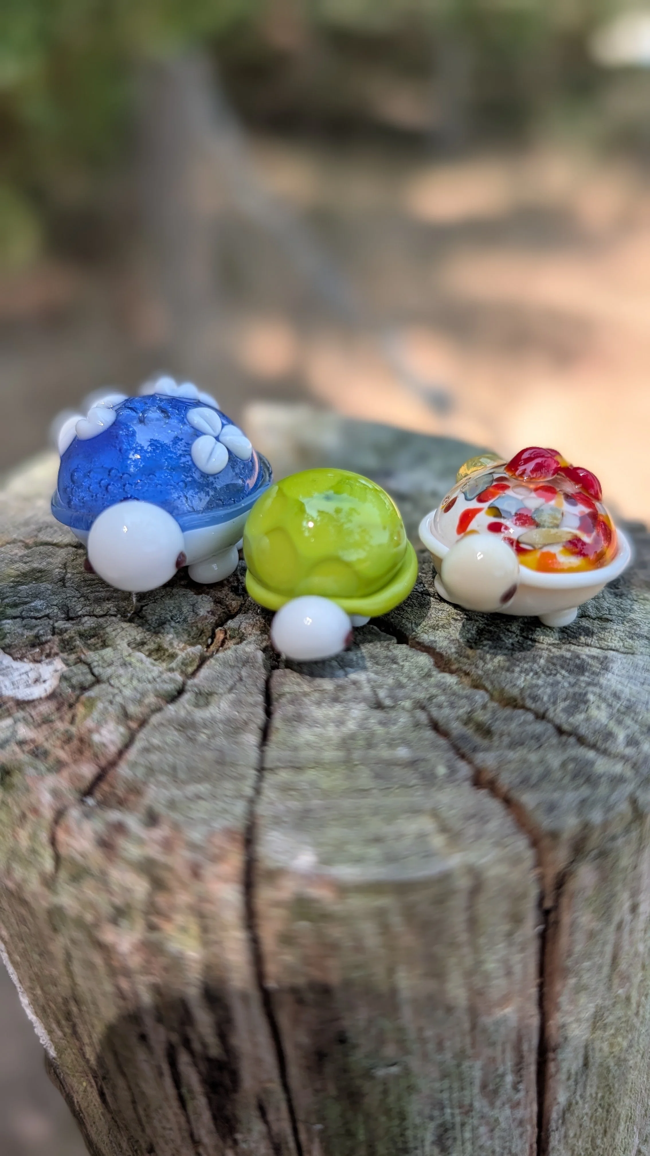 Trois petits objets en verre coloré sur une surface en bois, ils ressemblent à des oiseaux ou des animaux miniatures avec des détails décoratifs multicolores.