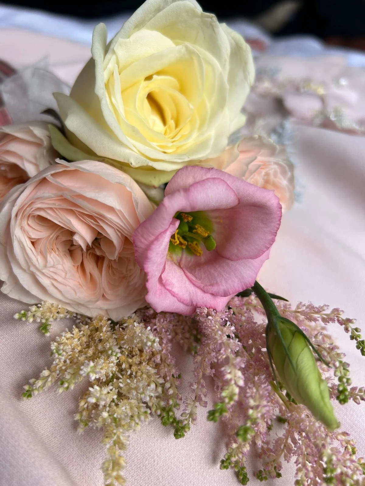 A close-up of a bouquet of flowers including a white rose, a pink rose, a pink lisianthus, and small white filler flowers on a soft pink fabric background.