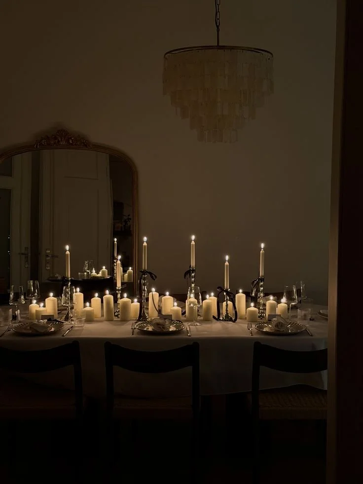 Dinner table set with candles, glasses, plates, and silverware in a dimly lit room with a chandelier overhead.
