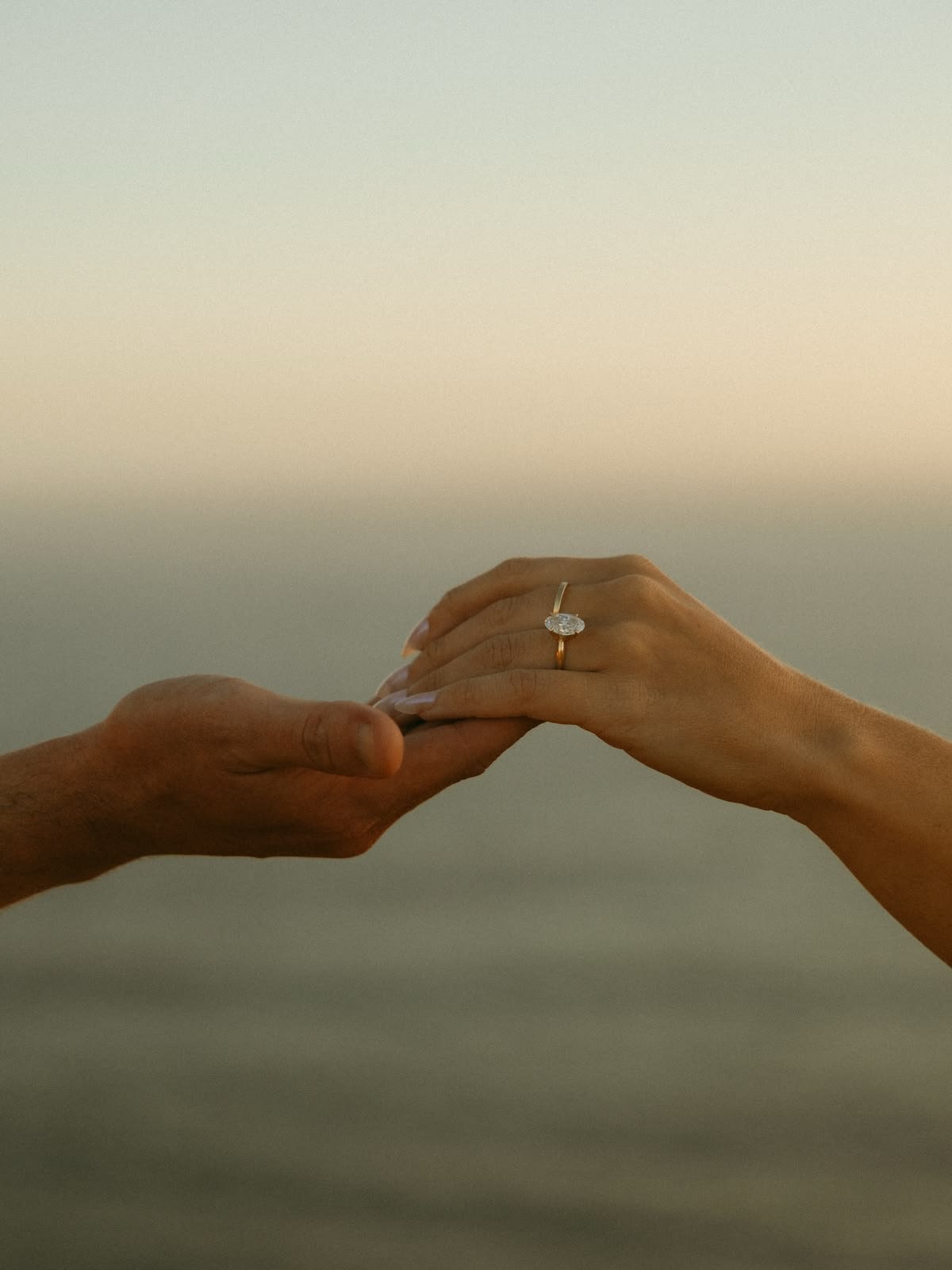 A person's hand gently holding another person's hand, which is wearing an engagement ring with a large oval diamond. The background is plain and neutral-colored.