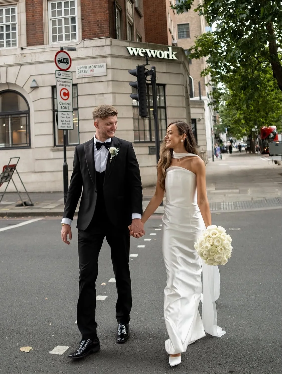 A newlywed couple in wedding attire holding hands and smiling while walking on a city street, with buildings, trees, and traffic lights in the background.