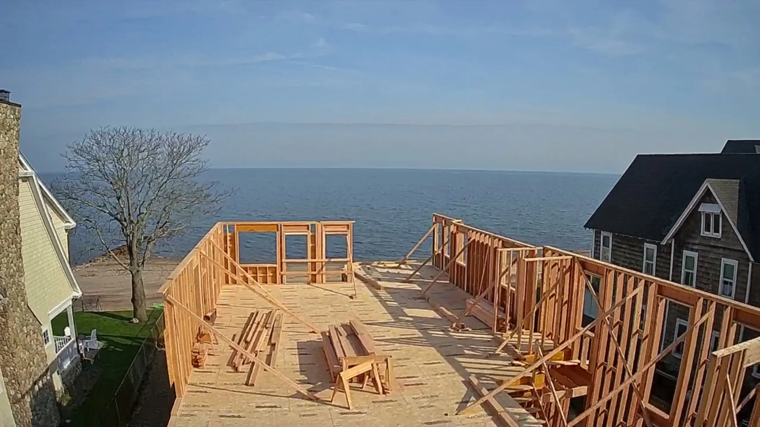 A house under construction with a view of the ocean or lake in the background. The wooden framing for a deck or porch is being built, and there are neighboring houses visible.