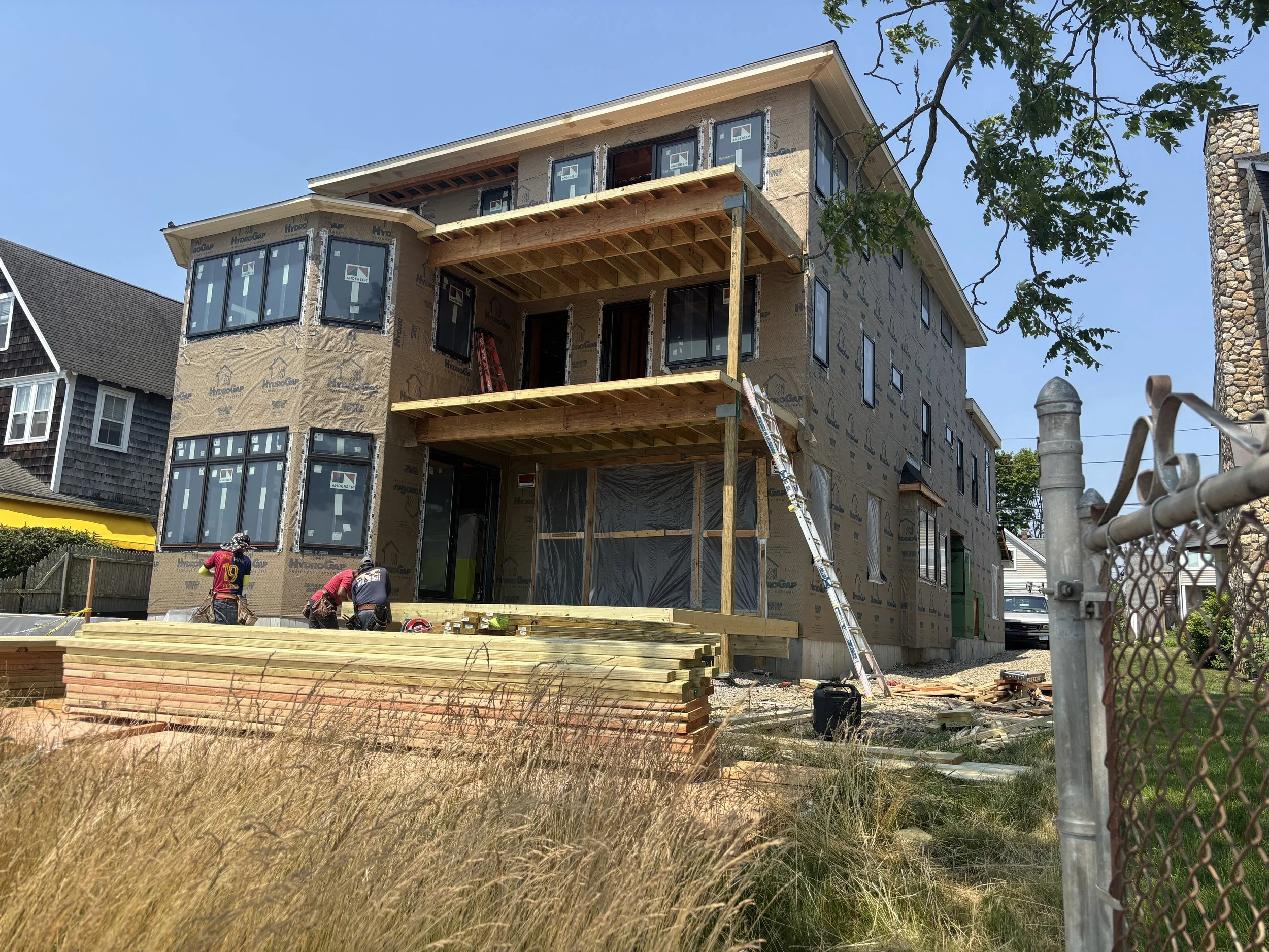 Construction workers are building a multi-story house, with the exterior walls covered in house wrap, and the framing for a balcony extending from the second floor. The house has large windows, and there's a ladder leaning against the balcony. The sc