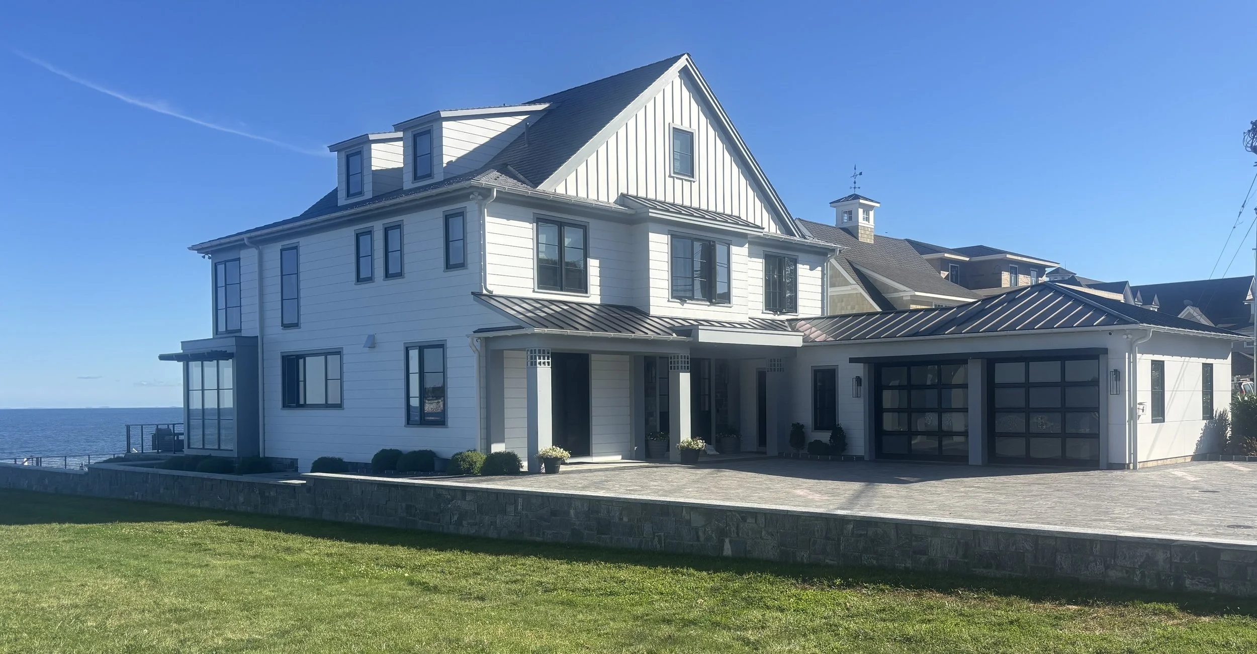 A large, modern white house with multiple levels, black window frames, and a garage, situated near water under a clear blue sky.