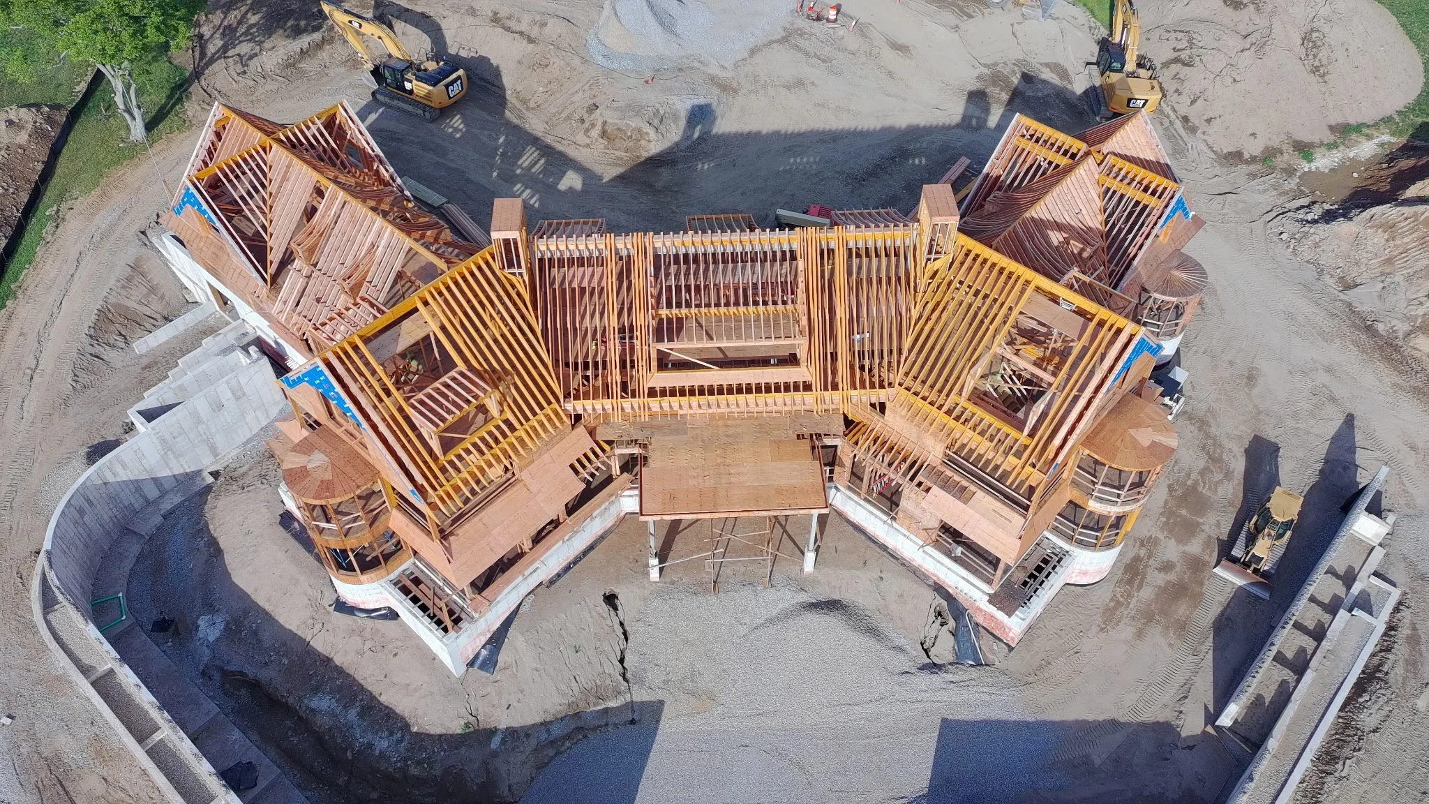 An aerial view of a house under construction with wooden framing, construction machinery, and surrounding dirt and concrete areas.
