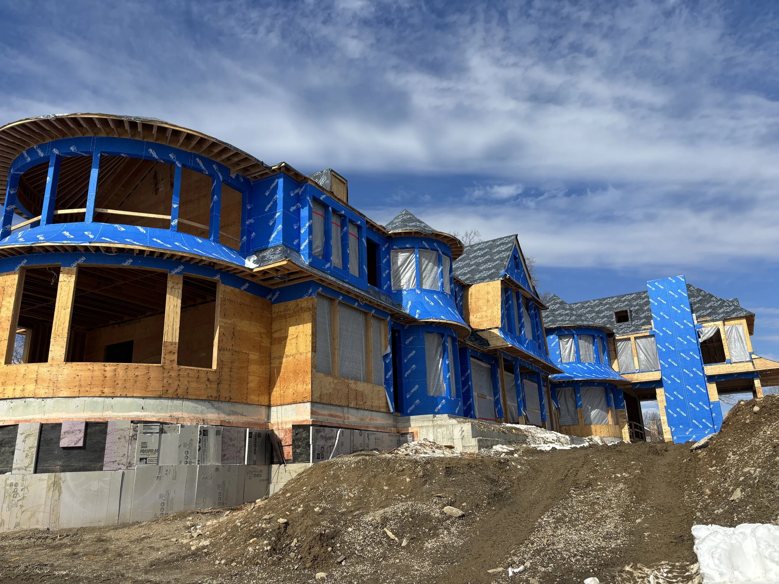 A house under construction with wooden framing and blue weather-resistant building wrap. The roof is partly finished, and the sky is partly cloudy with mountains in the background.