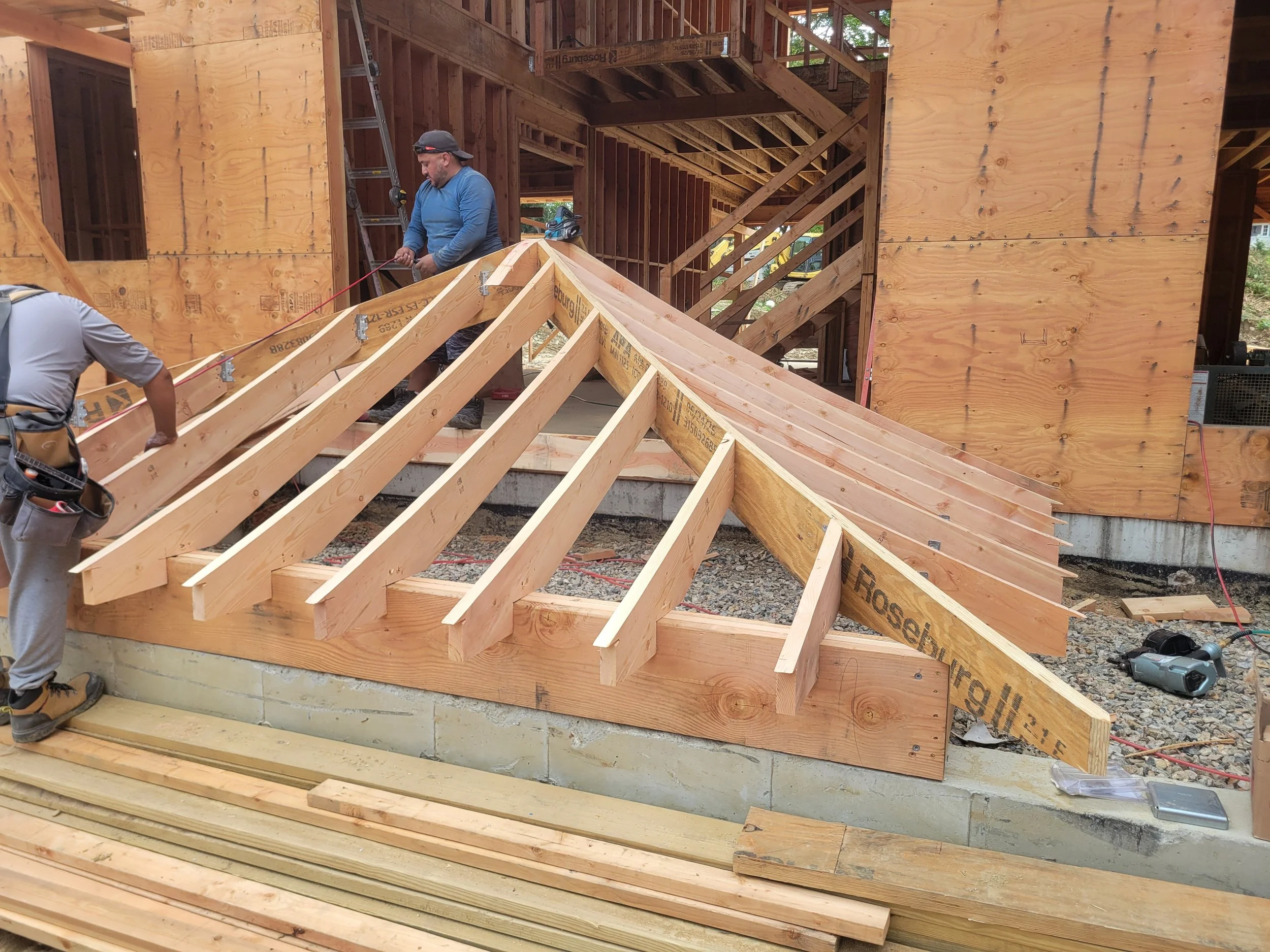 Construction workers building the wooden frame of a house. One man is working on the roof structure, while another is partially visible.