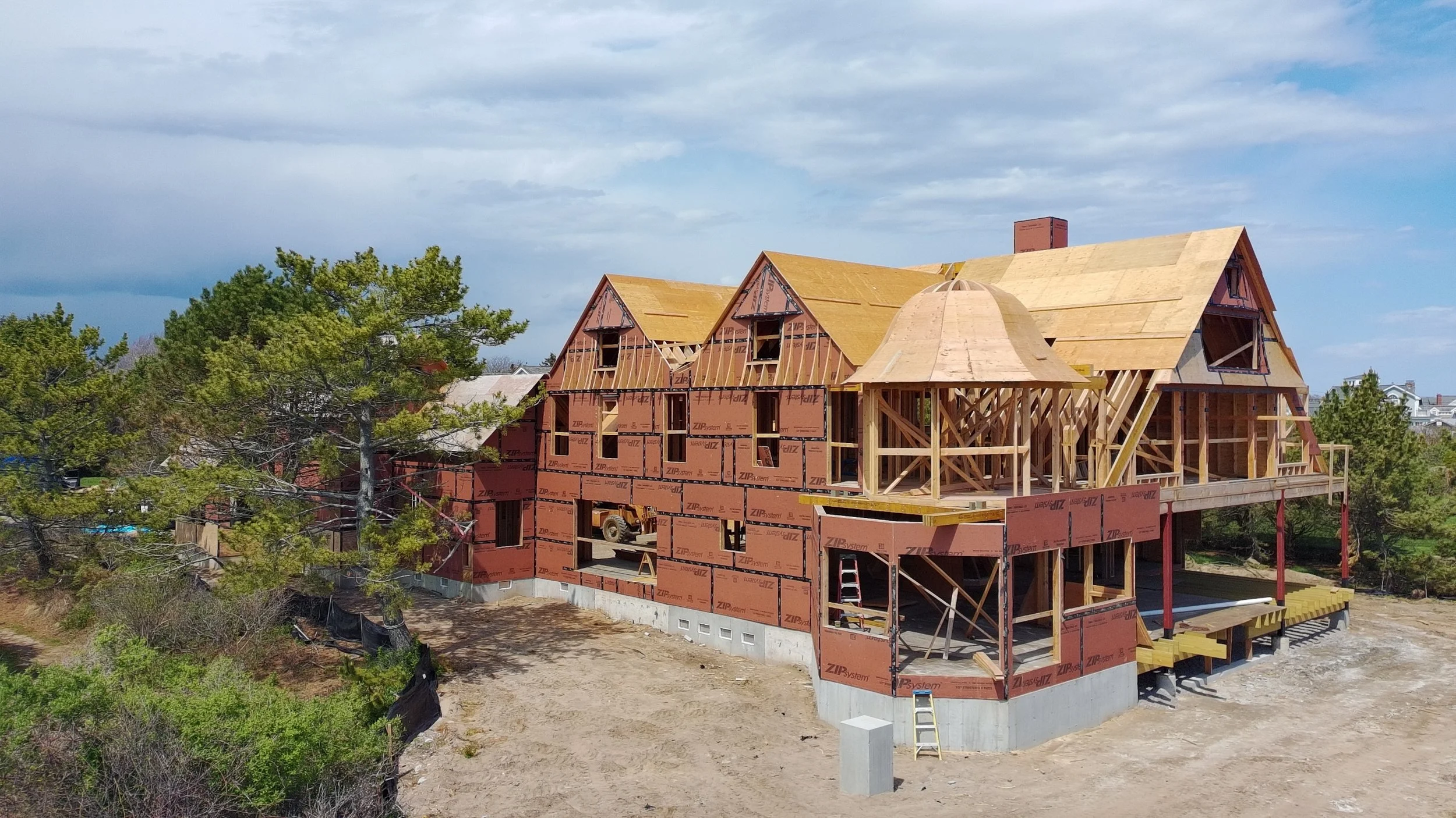 A house under construction with a wooden framework and partially installed roof, surrounded by trees and clear sky.