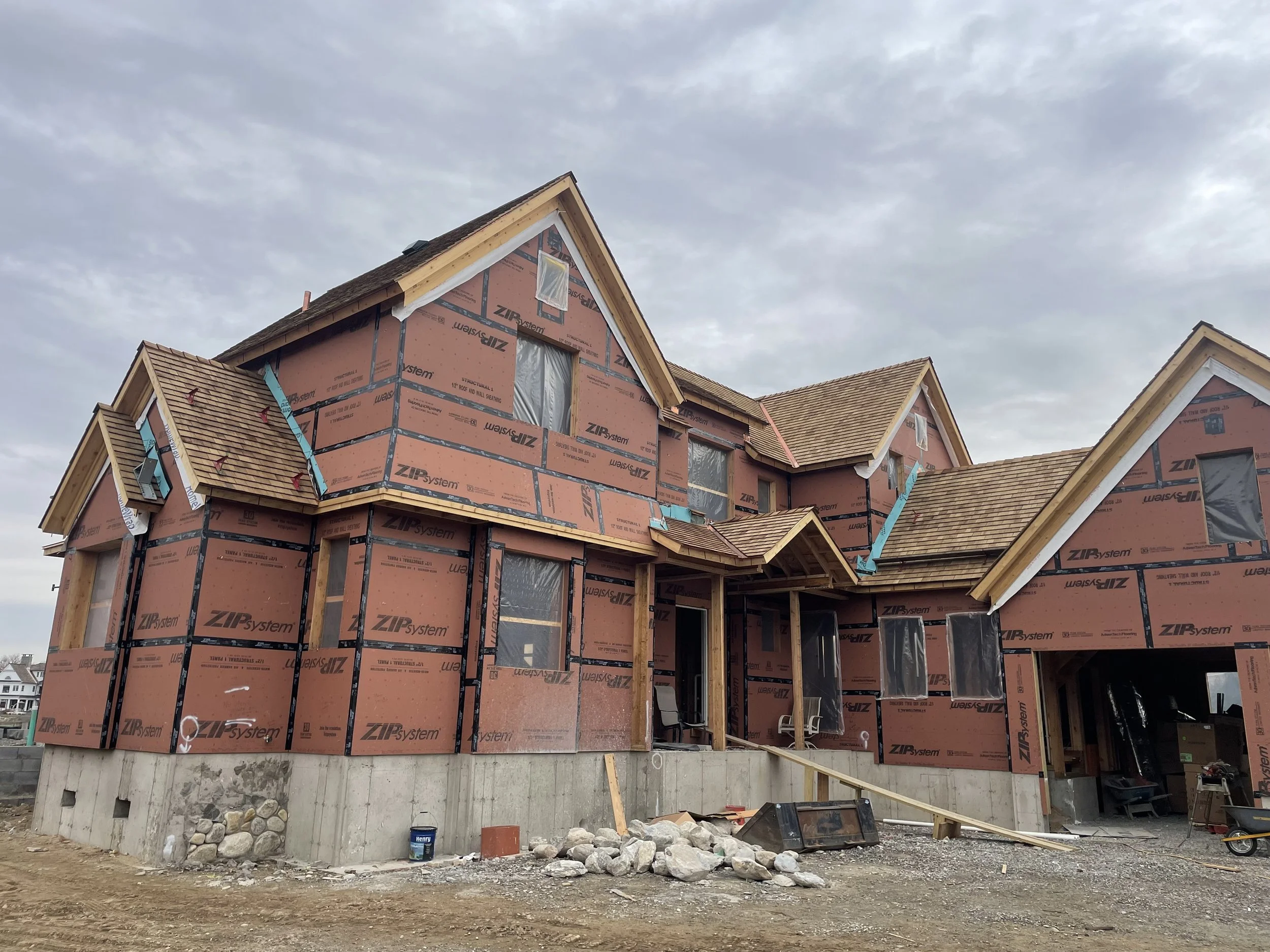 Under construction house with partial roofing and exposed siding, surrounded by construction materials and debris, with a cloudy sky in the background.