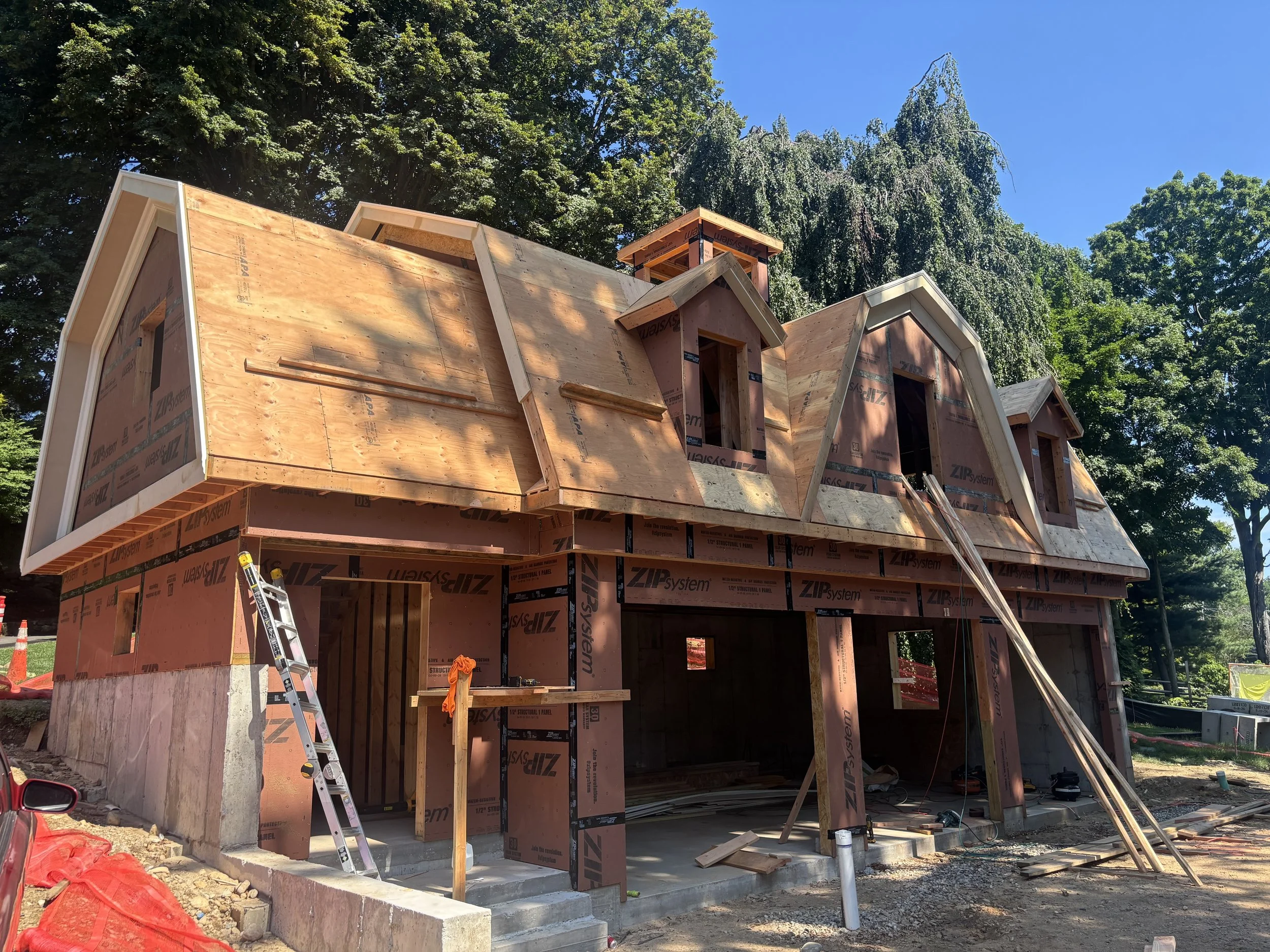 A house under construction with wooden framing and a partially completed roof, set against a background of tall green trees and a blue sky.