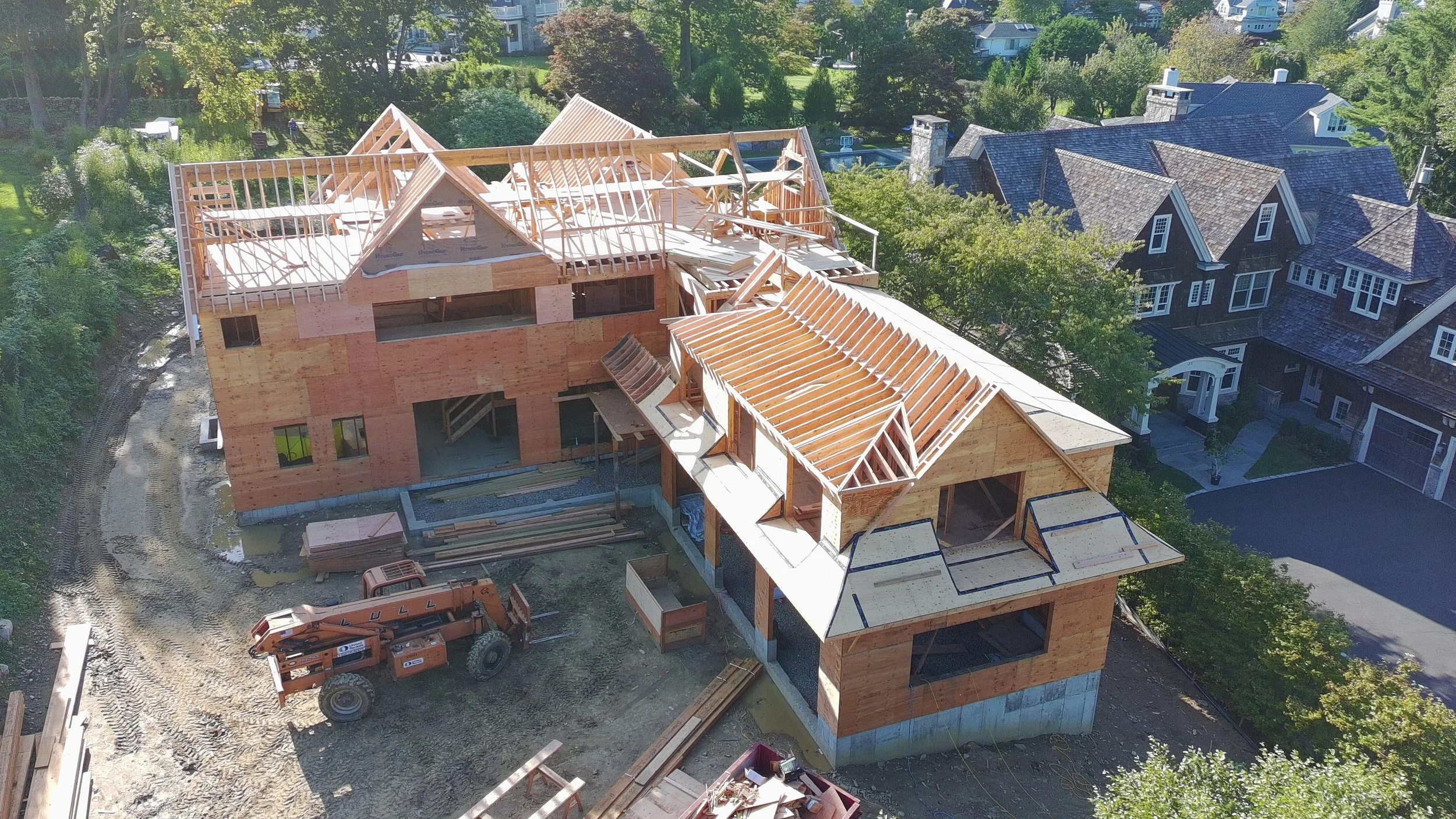 A house under construction with a wooden frame and roof trusses, surrounded by trees and neighboring houses.