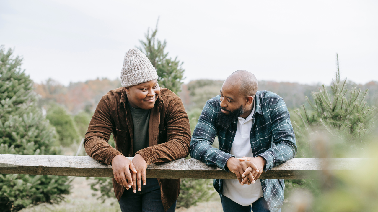 Two men lean on a wooden fence outdoors, engaging in conversation, surrounded by pine trees and a natural landscape.