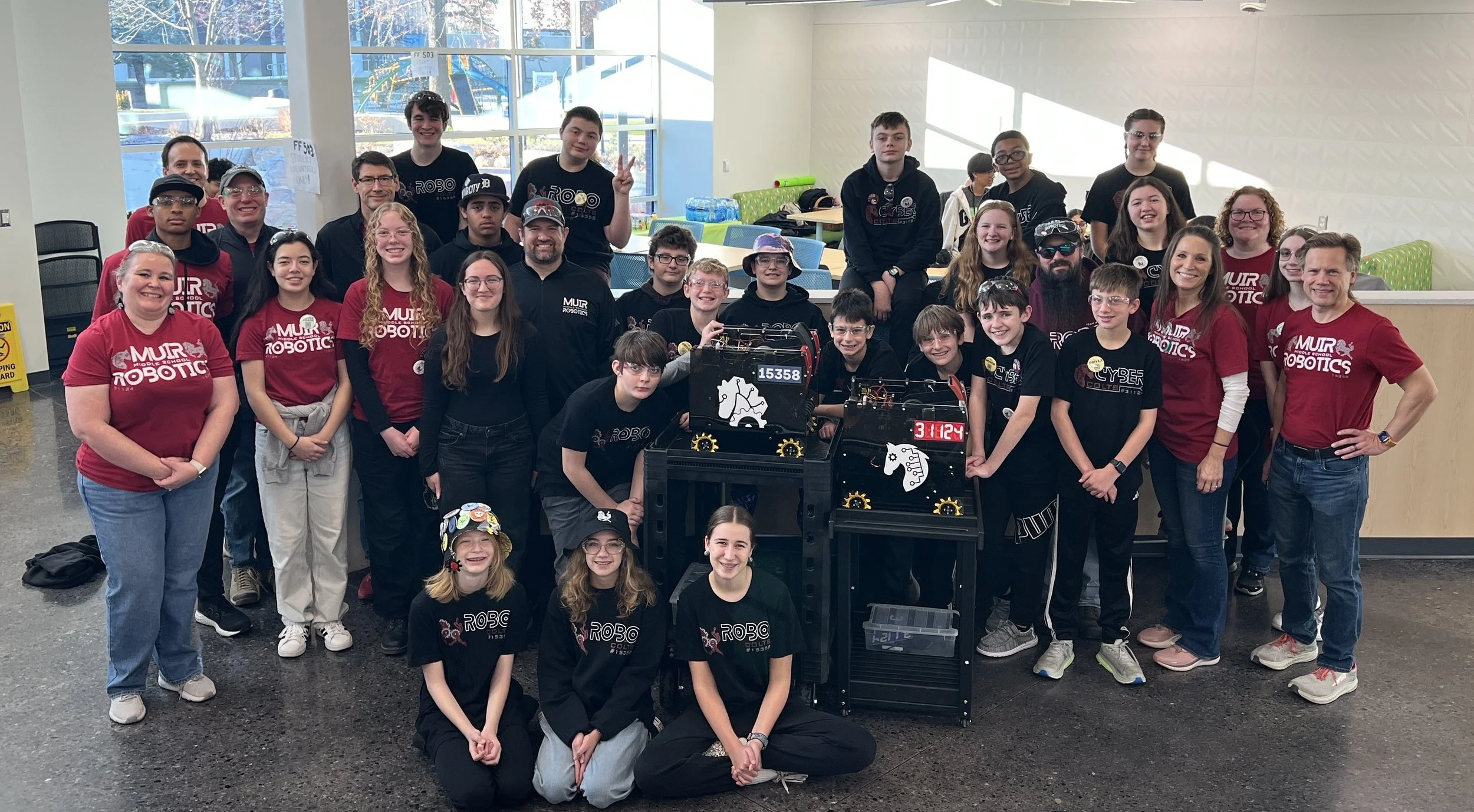 A group of students and adults gathered indoors around robot projects, with some there to show off their robots at a robotics competition or event.