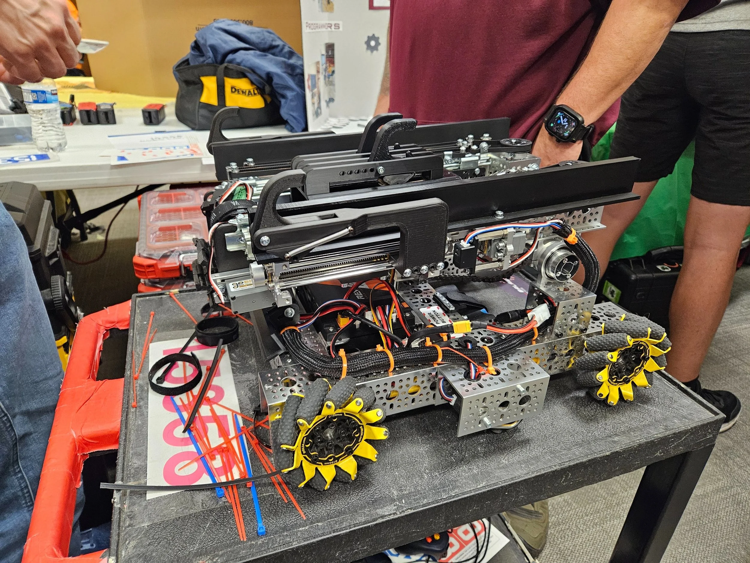A person working on a robot at a table with various tools and parts, including yellow wheels, wires, and metal structures, in a workshop or event setting.
