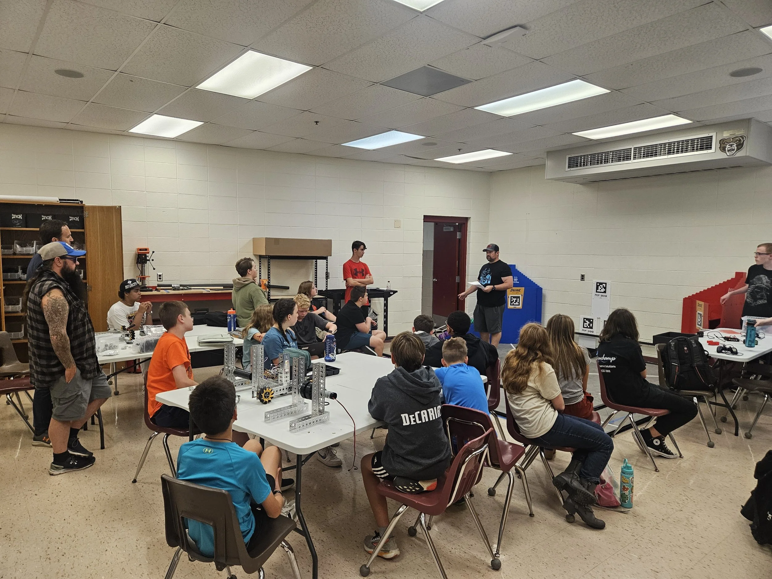 A group of young people gathered in a classroom or workshop setting, some sitting and some standing, with a man in a black shirt and cap speaking to the group. The room has a white cinderblock wall, fluorescent ceiling lights, a red door, and shelves with various items. A table with mechanical or robotic parts is in the foreground.