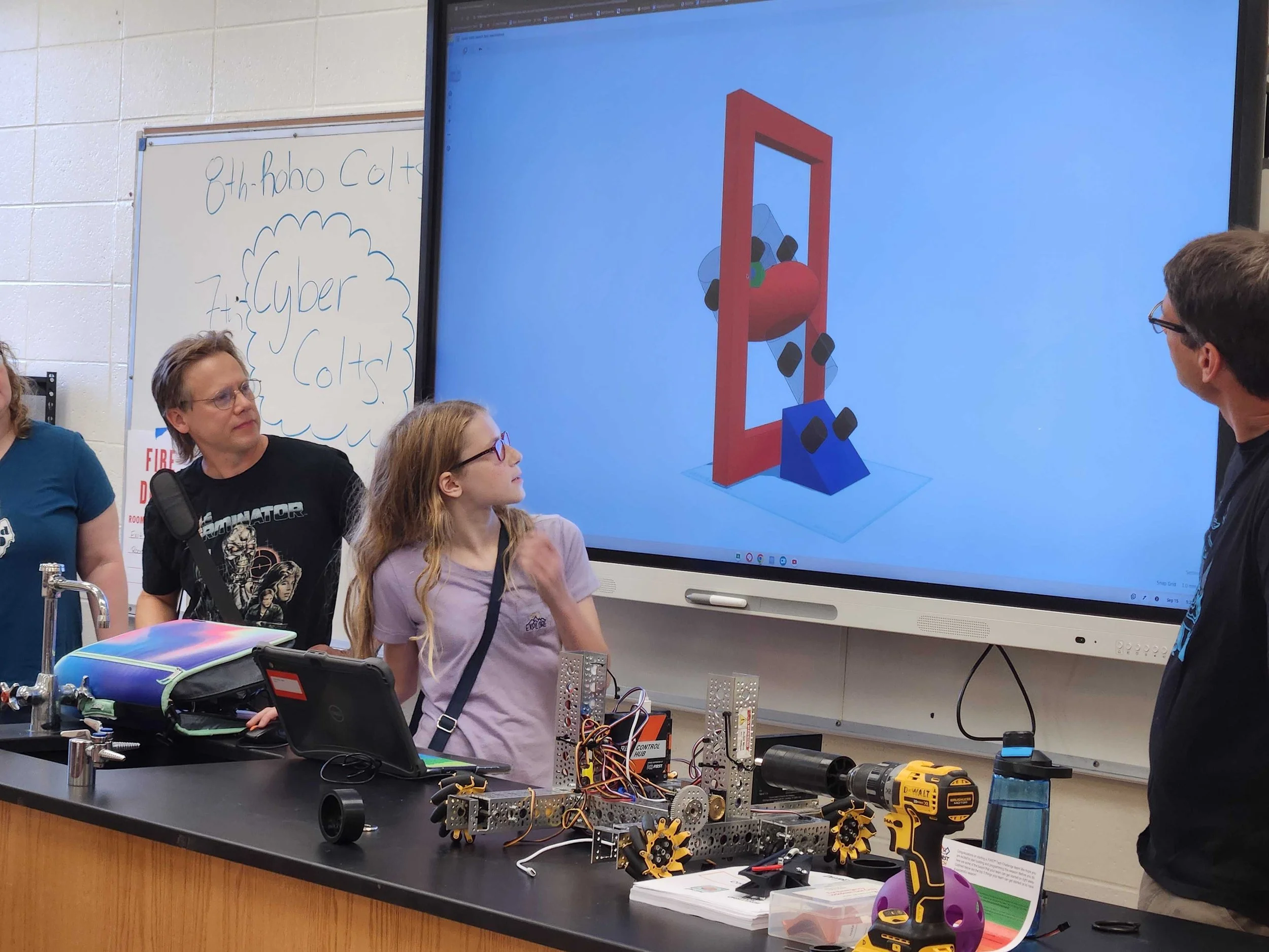 Students in a classroom working on robotics and engineering projects with a large screen displaying a 3D model of a robot or machine part.