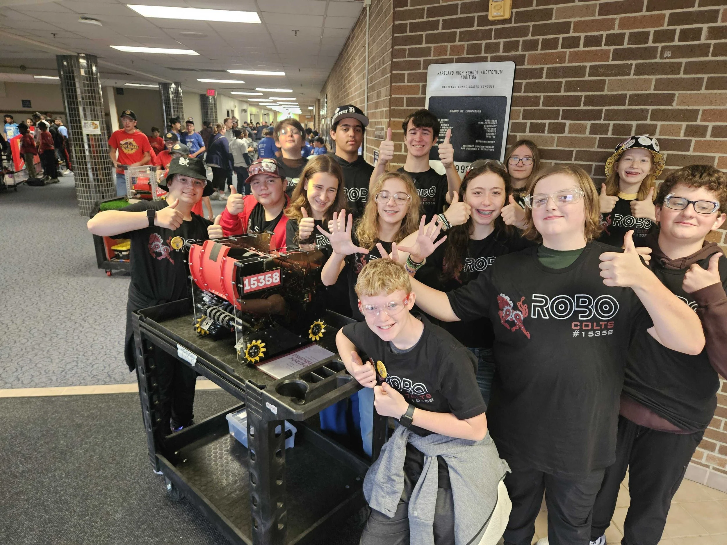 A group of students assembled around a robot on a cart, giving thumbs up and smiling in a school hallway. The students are wearing black T-shirts with the word 'ROBO' and a robot logo. There are many others in the background, some wearing hats and safety glasses.