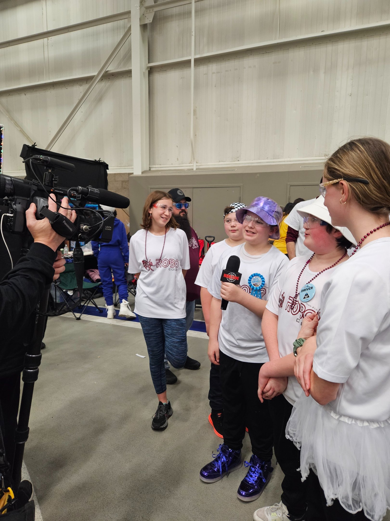 A group of children and adults being interviewed by a news crew inside a large indoor space, possibly a warehouse or convention center. The children are wearing various costumes, some with robes, ribbons, and large boots, and are smiling while talking to the interviewer.