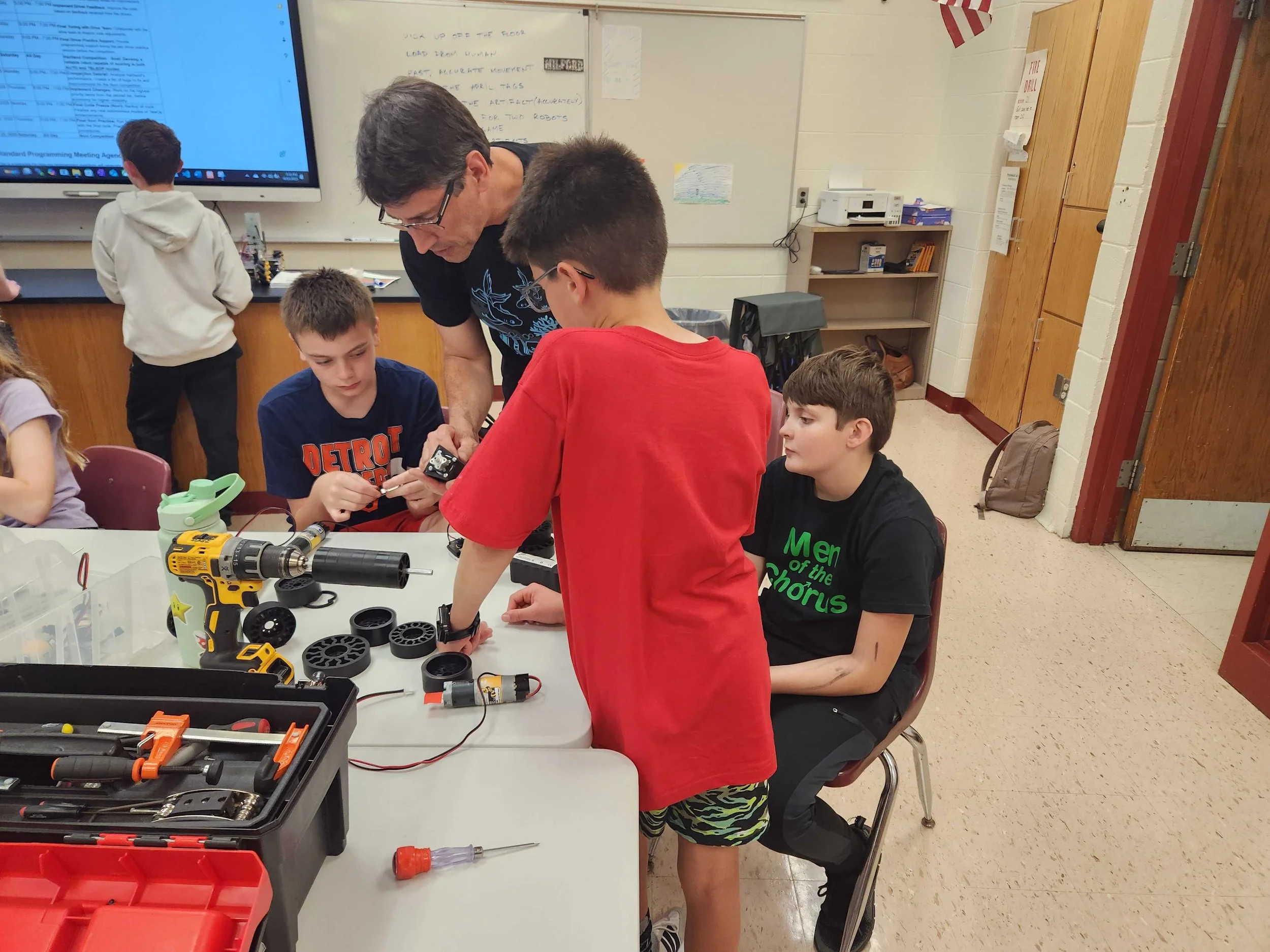 A classroom scene with a teacher and students working on robotics projects. The teacher is showing a robot to a group of boys sitting and standing around the table. Various tools and robot parts are on the table, with a large screen displaying information in the background.